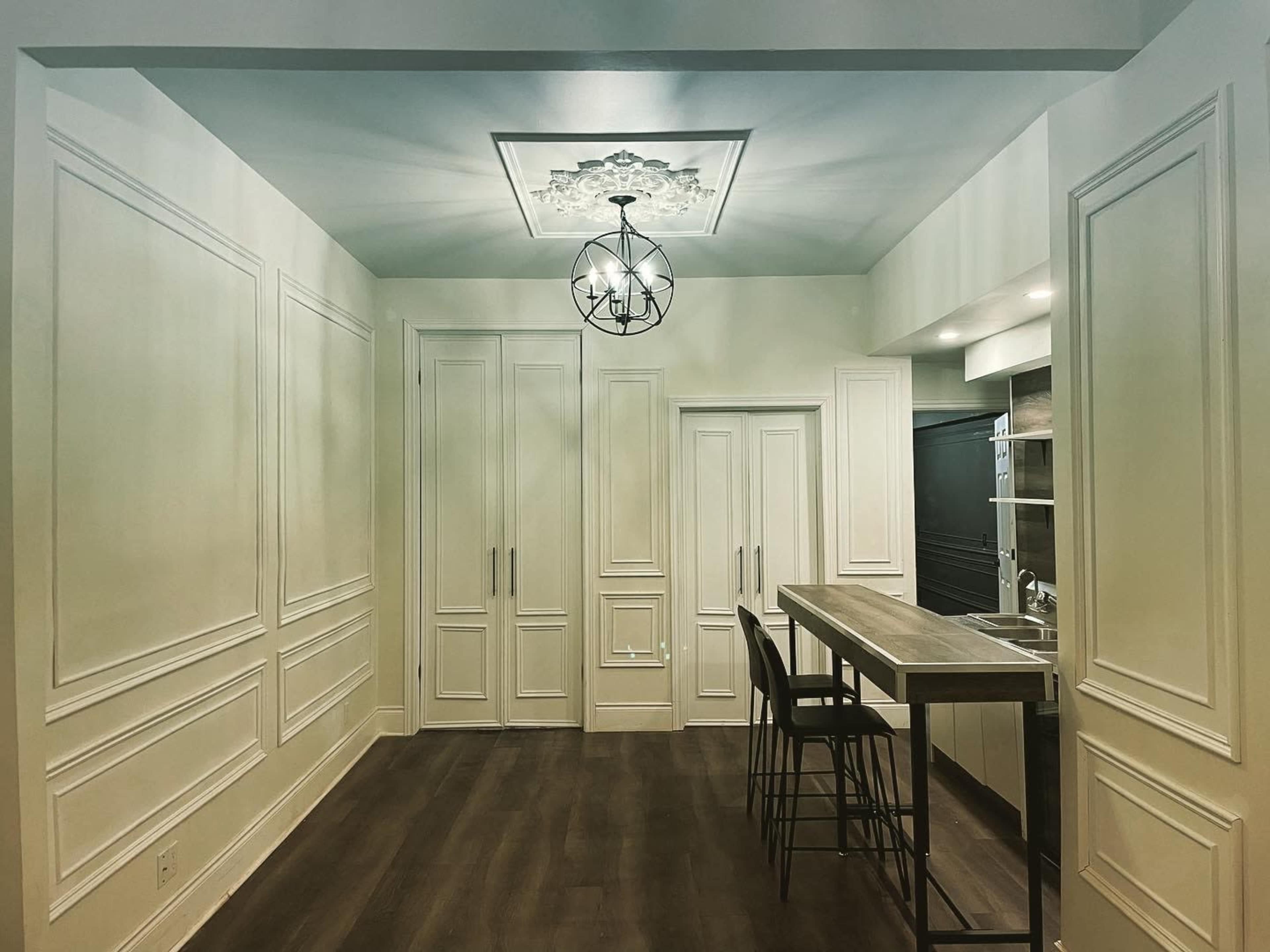 A modern kitchen area features a central wooden bar table with high stools, flanked by light-colored cabinetry and a decorative ceiling fixture.