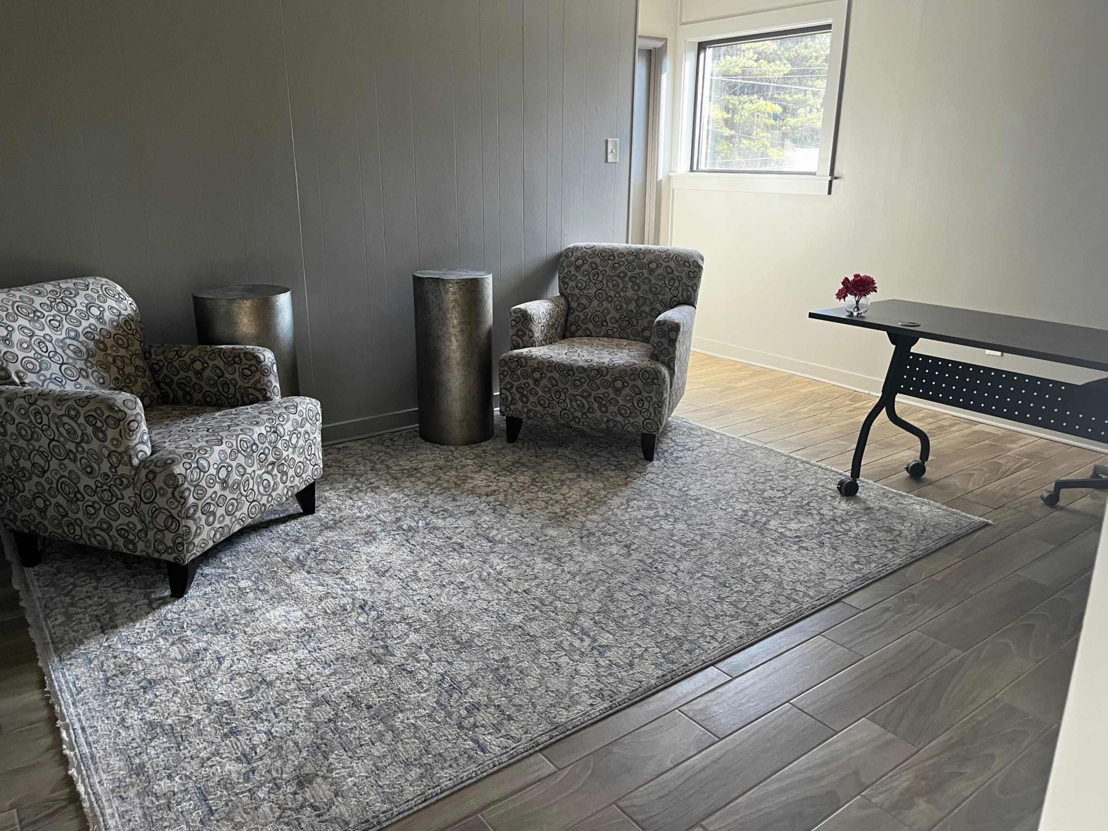 The image shows a small, modern room with two patterned chairs, a metallic cylindrical table, and a small desk on a wooden floor.