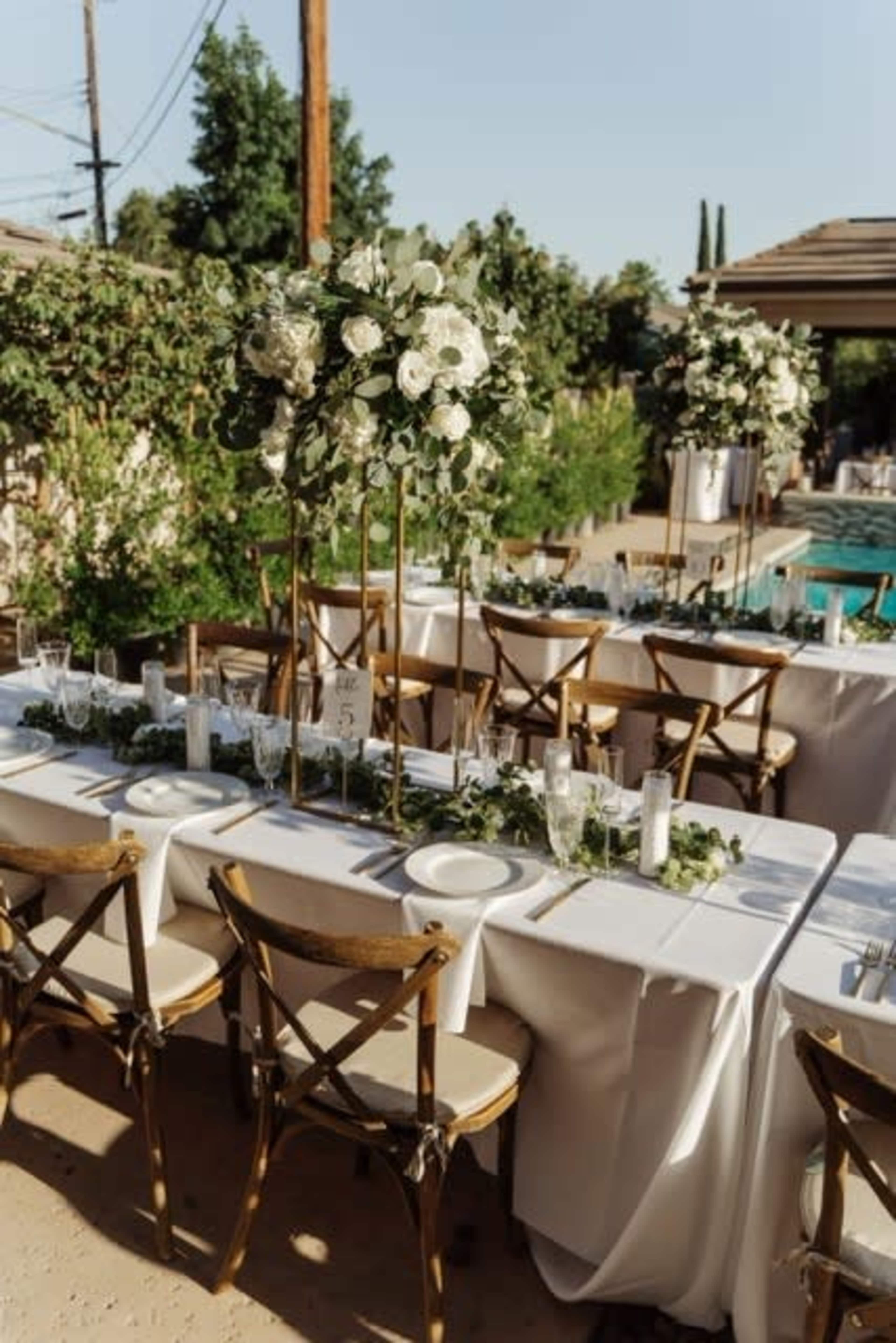 A set of elegantly arranged dining tables with tall floral centerpieces is displayed outdoors beside a pool.