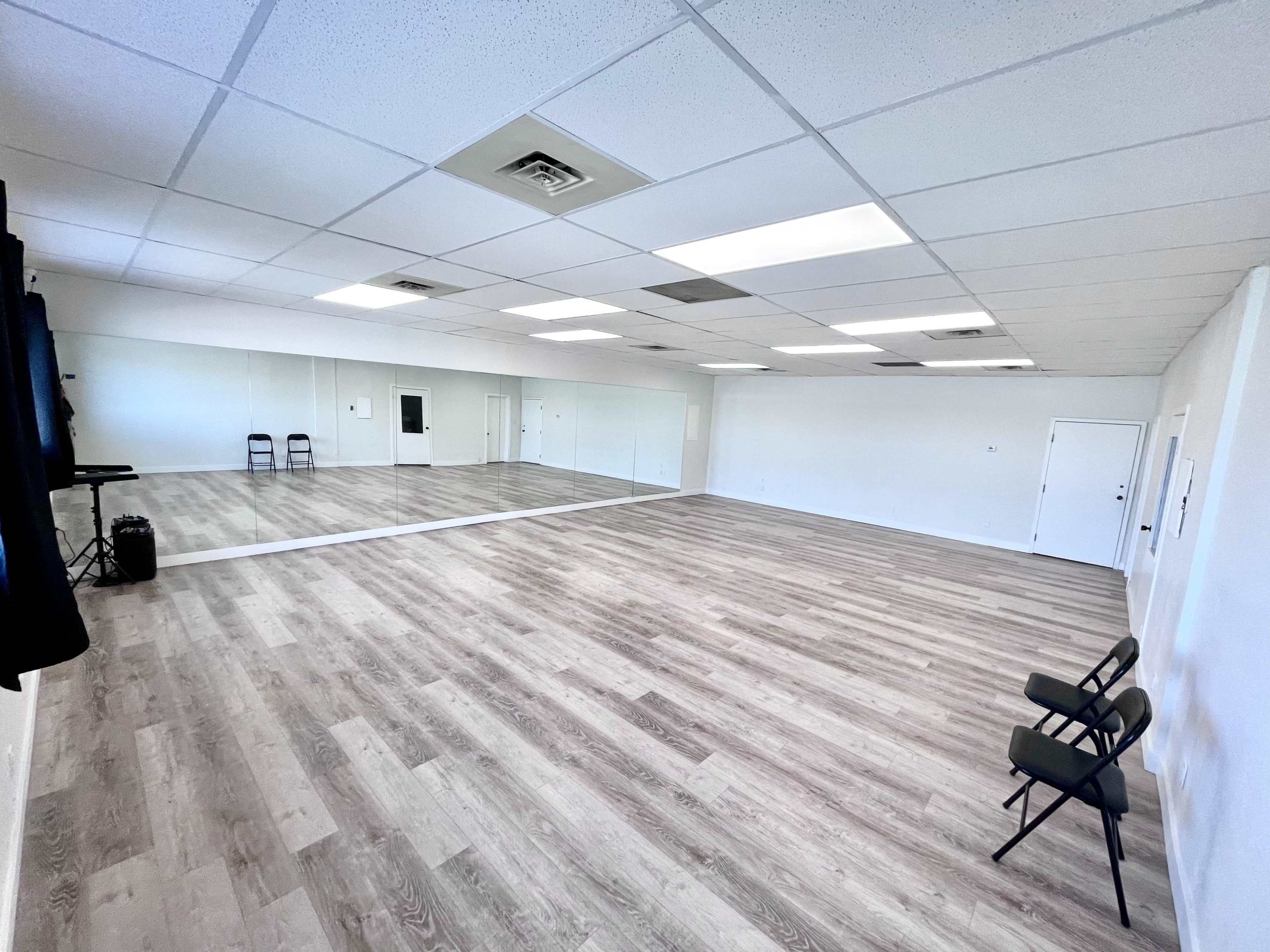 A spacious, empty room with wooden flooring, large mirrors on one wall, and two chairs positioned beside the wall.