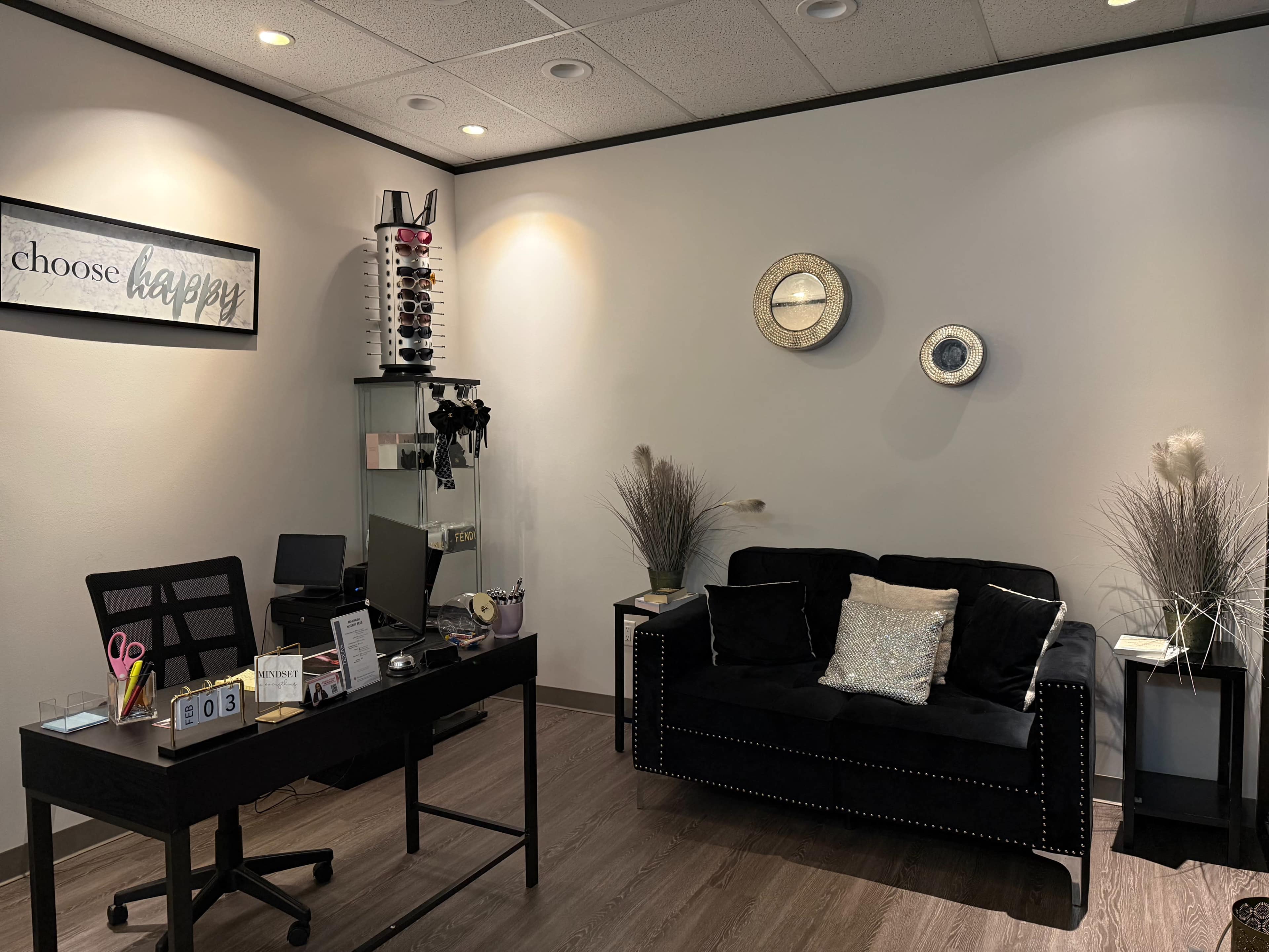 The image shows a modern office space featuring a black desk, a black sofa with a decorative pillow, a wall-mounted clock, and a display of eyeglasses on the wall.