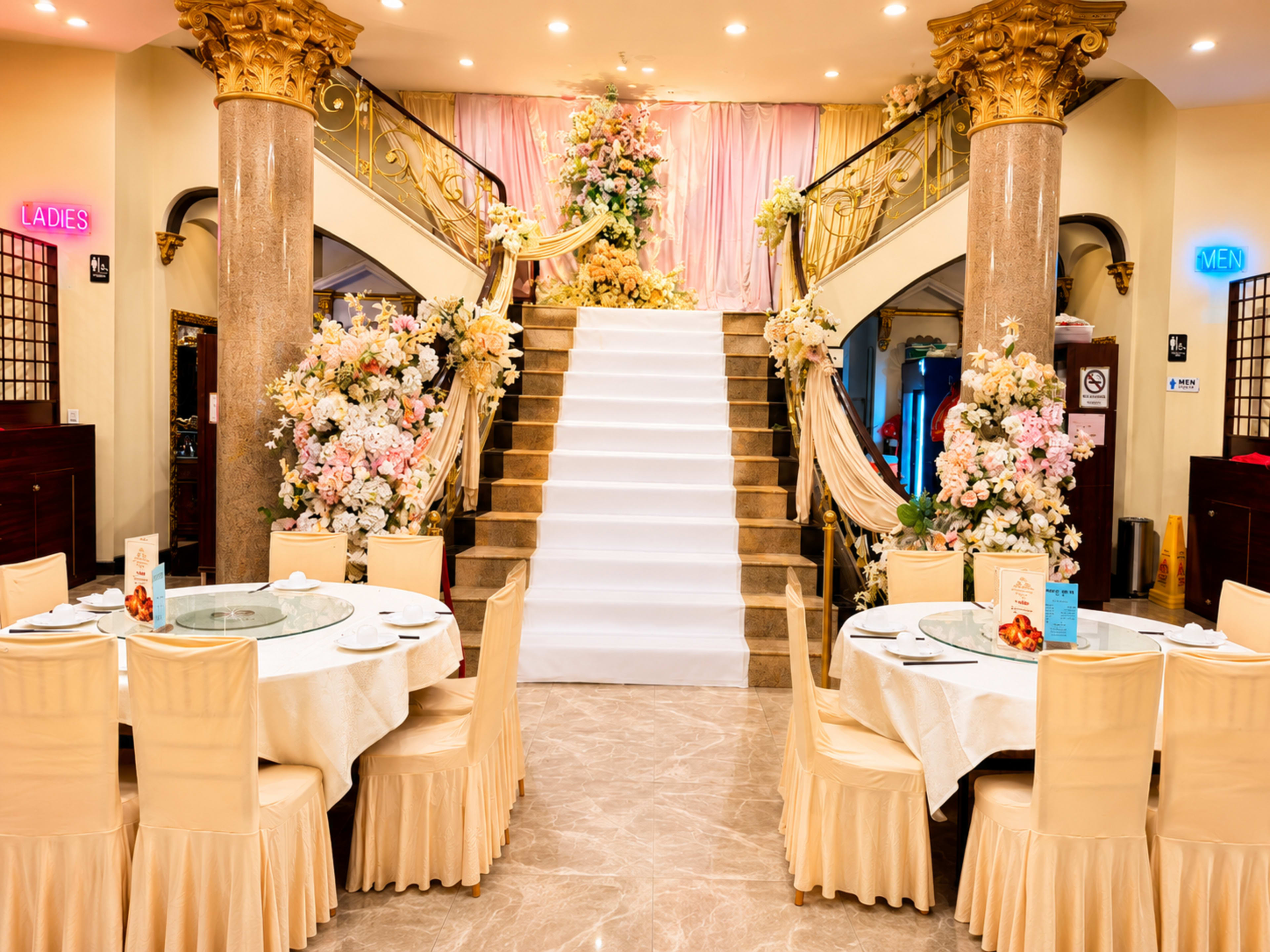 The image shows an elegantly decorated banquet hall with a grand staircase adorned with flowers, flanked by signs indicating "LADIES" and "MEN," and tables set for dining.