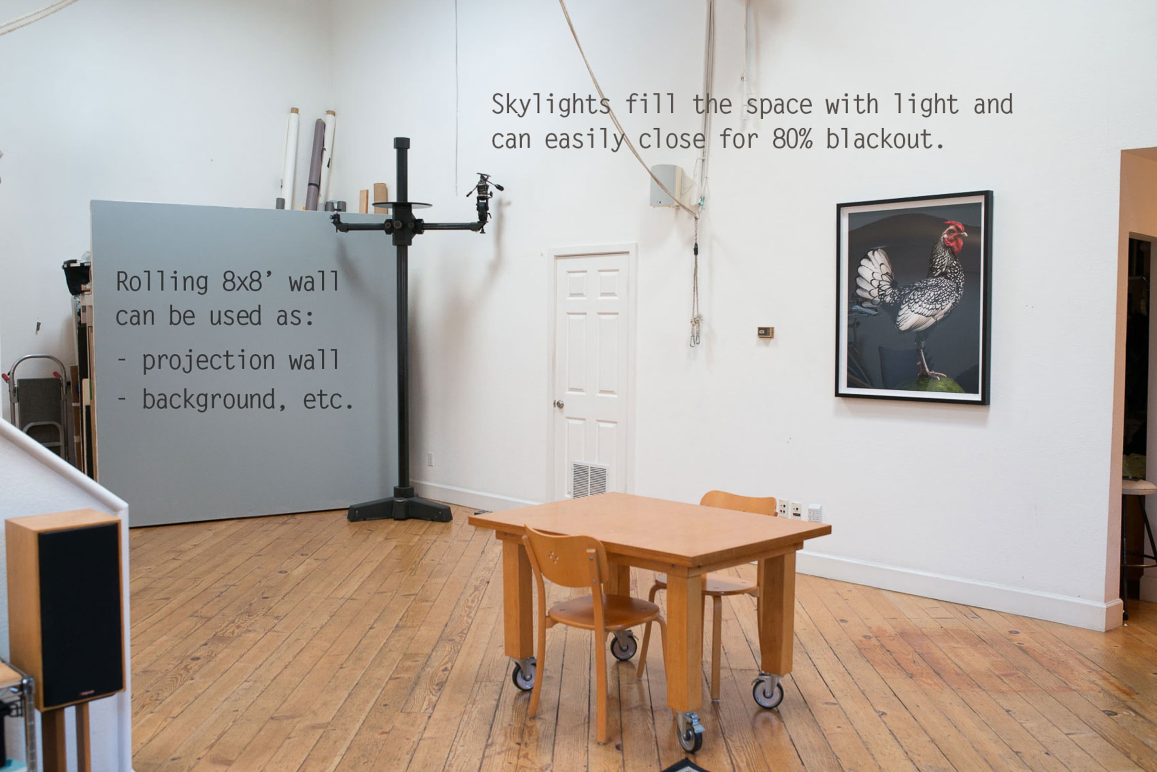 The image shows a well-lit studio space with skylights, featuring a wooden floor, a table with two chairs, and a large black-and-white photograph of a chicken on the wall.