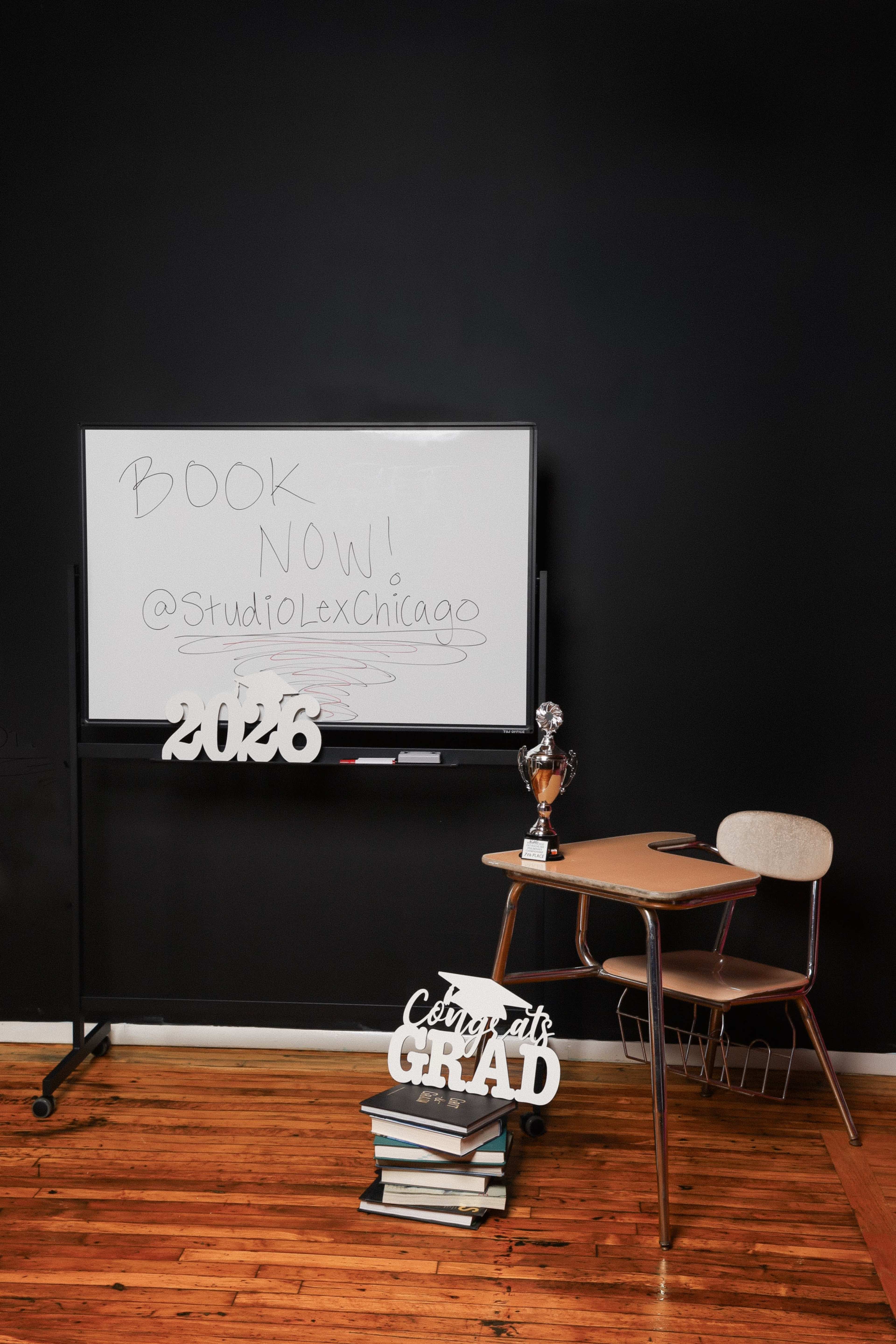 The image shows a classroom setup featuring a whiteboard with a message, a trophy on a desk, a stack of books, and decorations that read "Congrats Grad" and "2026," all against a black wall.