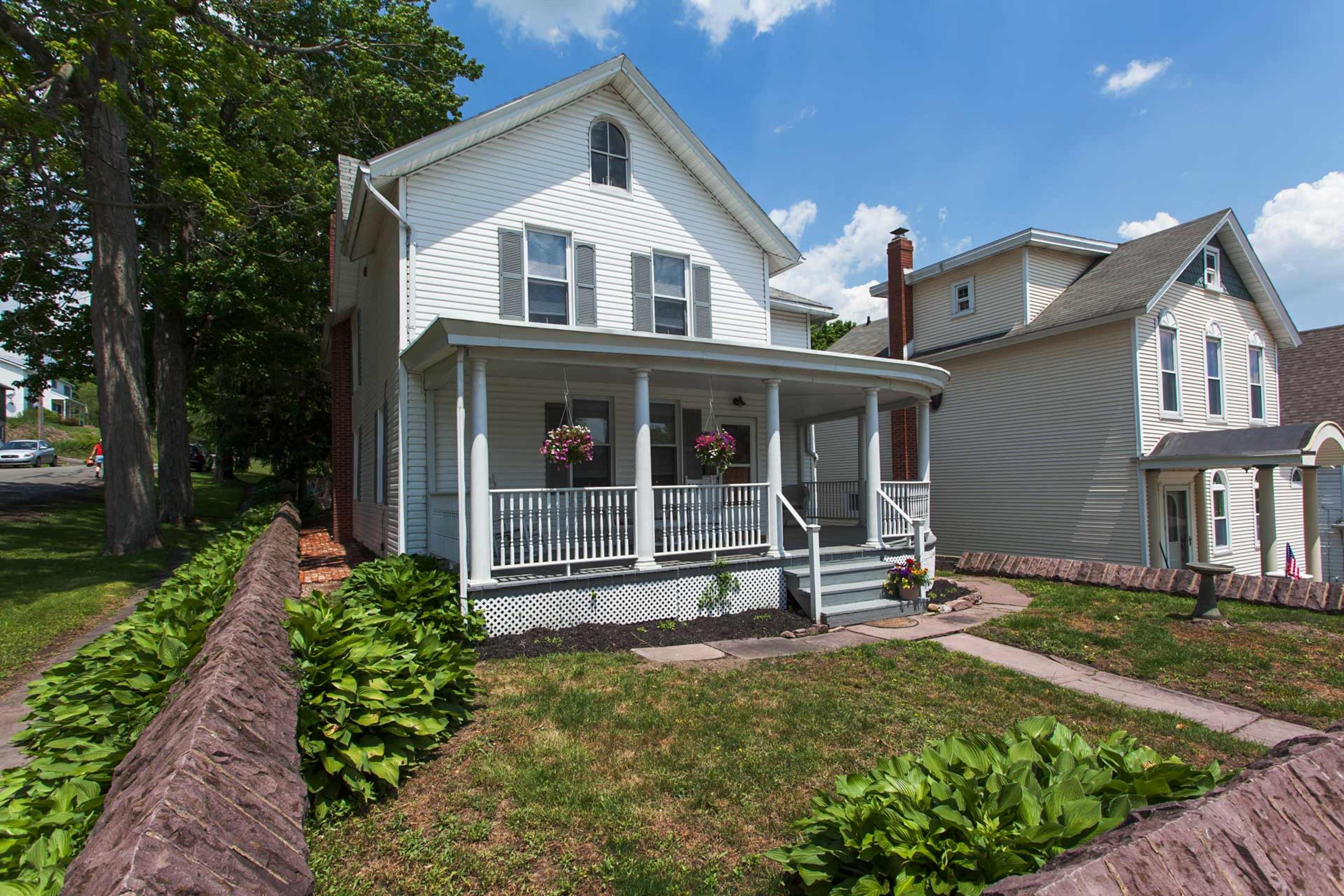 A two-story white house with a front porch, surrounded by a grassy lawn and a low stone wall.