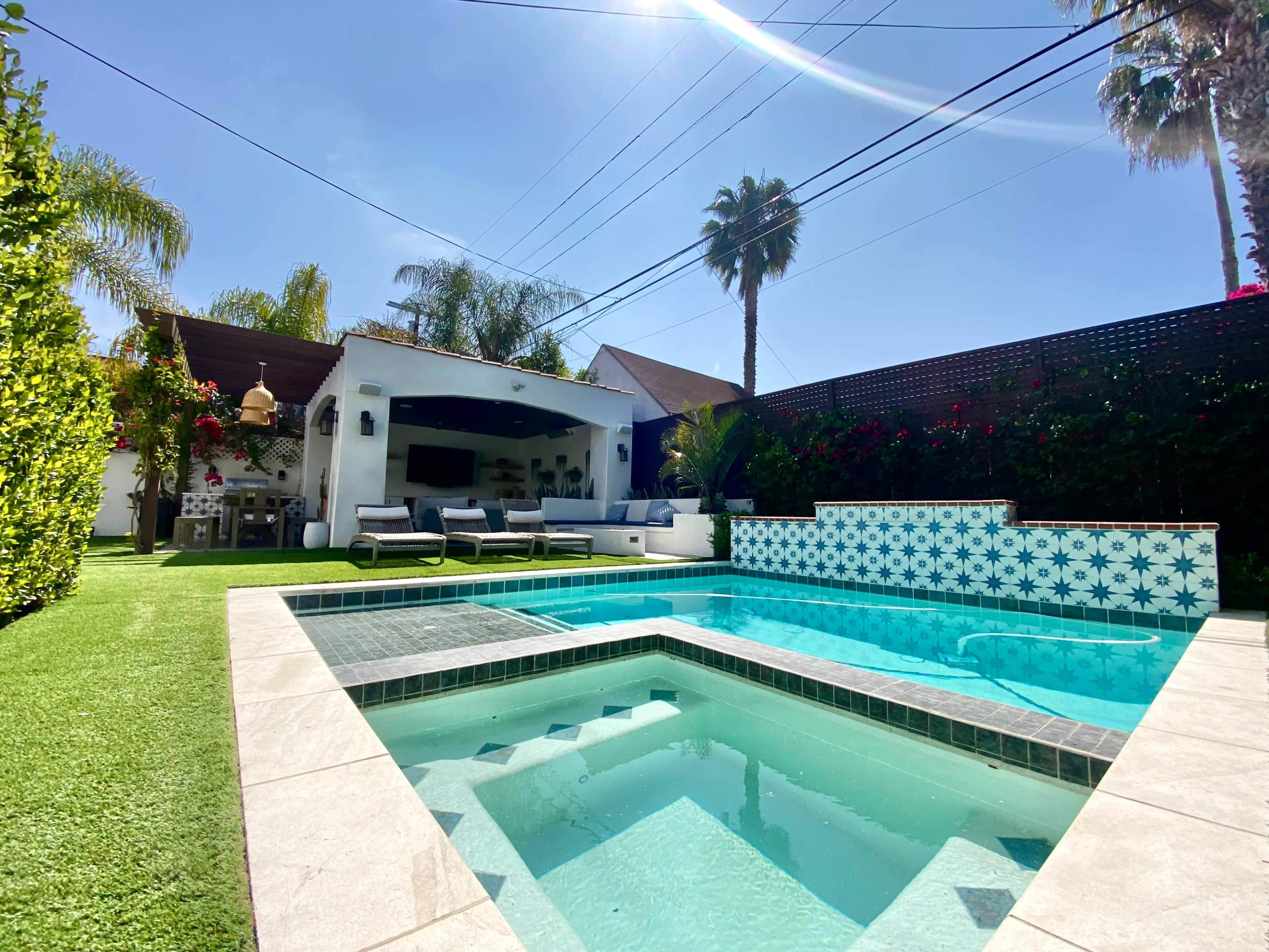The image shows a backyard with a swimming pool featuring tile designs, lounge chairs, and a house partially visible behind it, surrounded by palm trees and greenery.