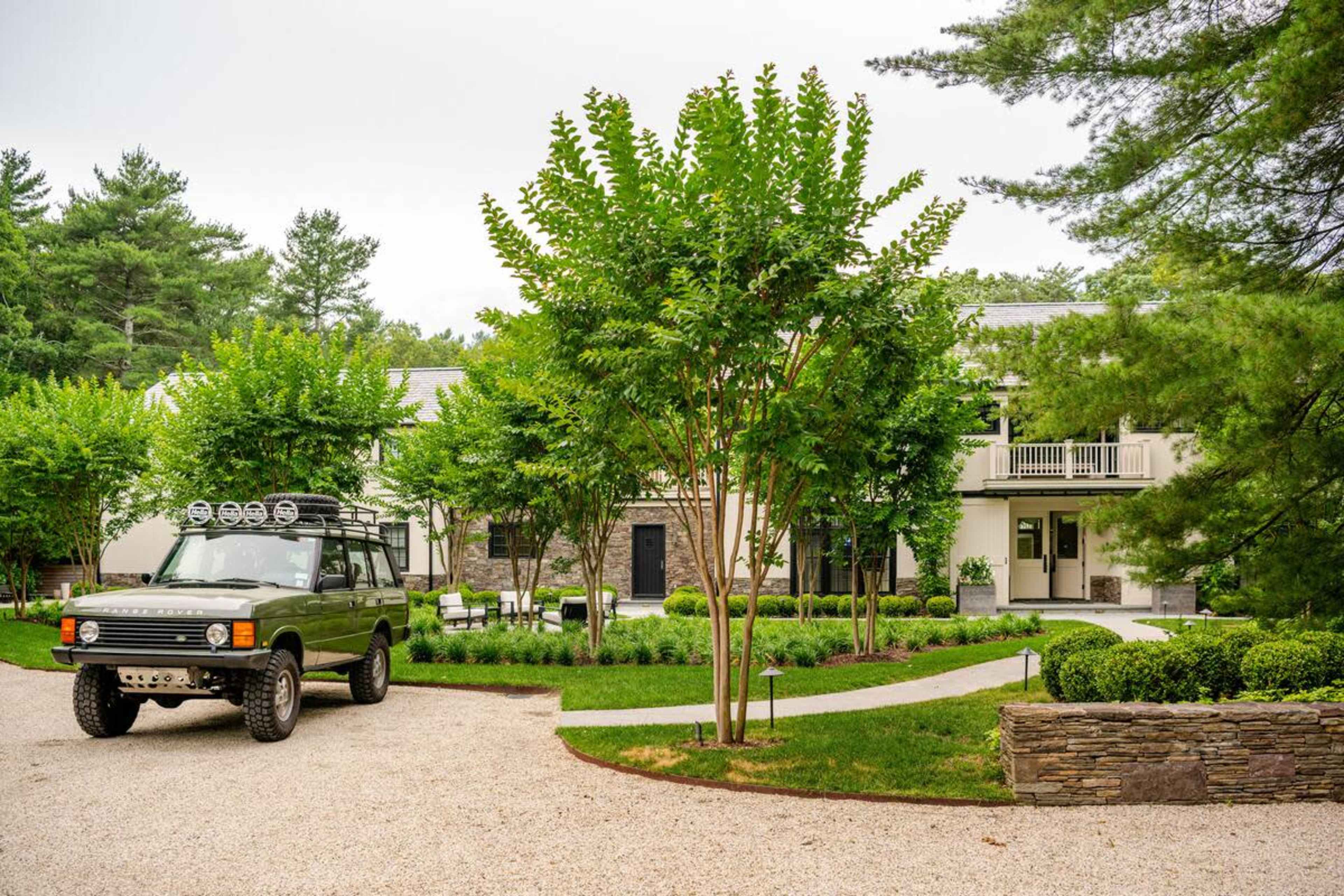 A large SUV is parked on a gravel driveway beside a landscaped yard with neatly trimmed shrubs and trees, leading up to a two-story house.