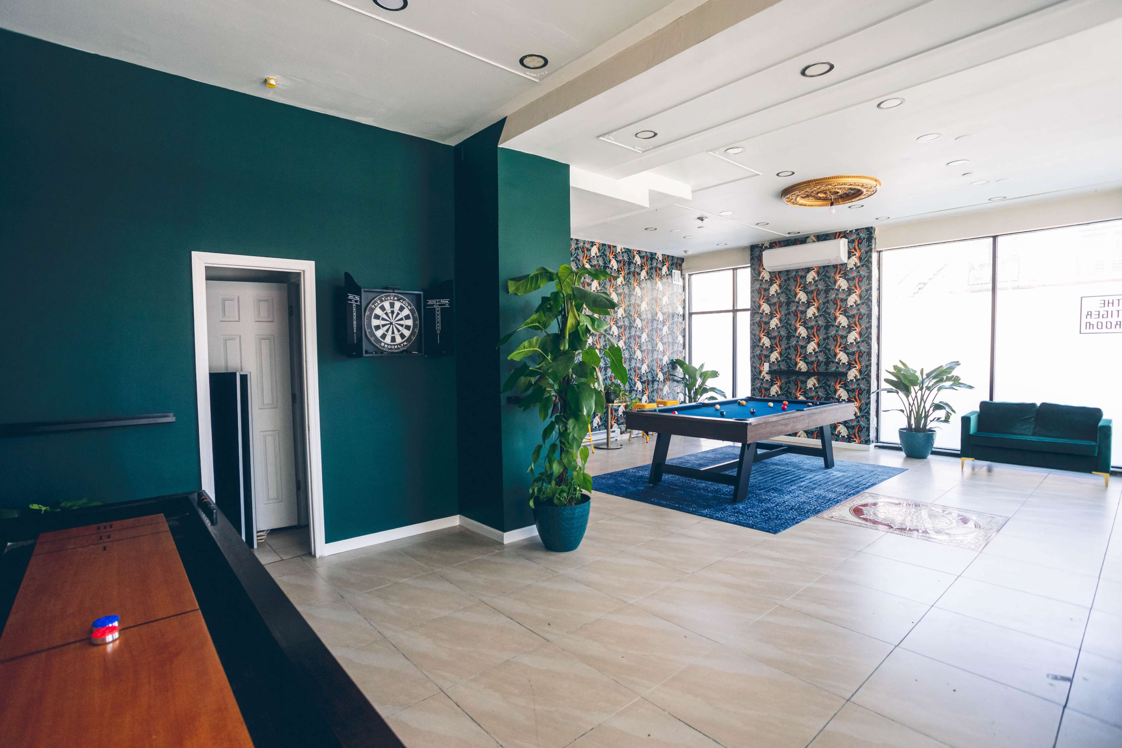 A recreational room with a pool table, a dartboard, and seating surrounded by indoor plants and decorative wallpaper.