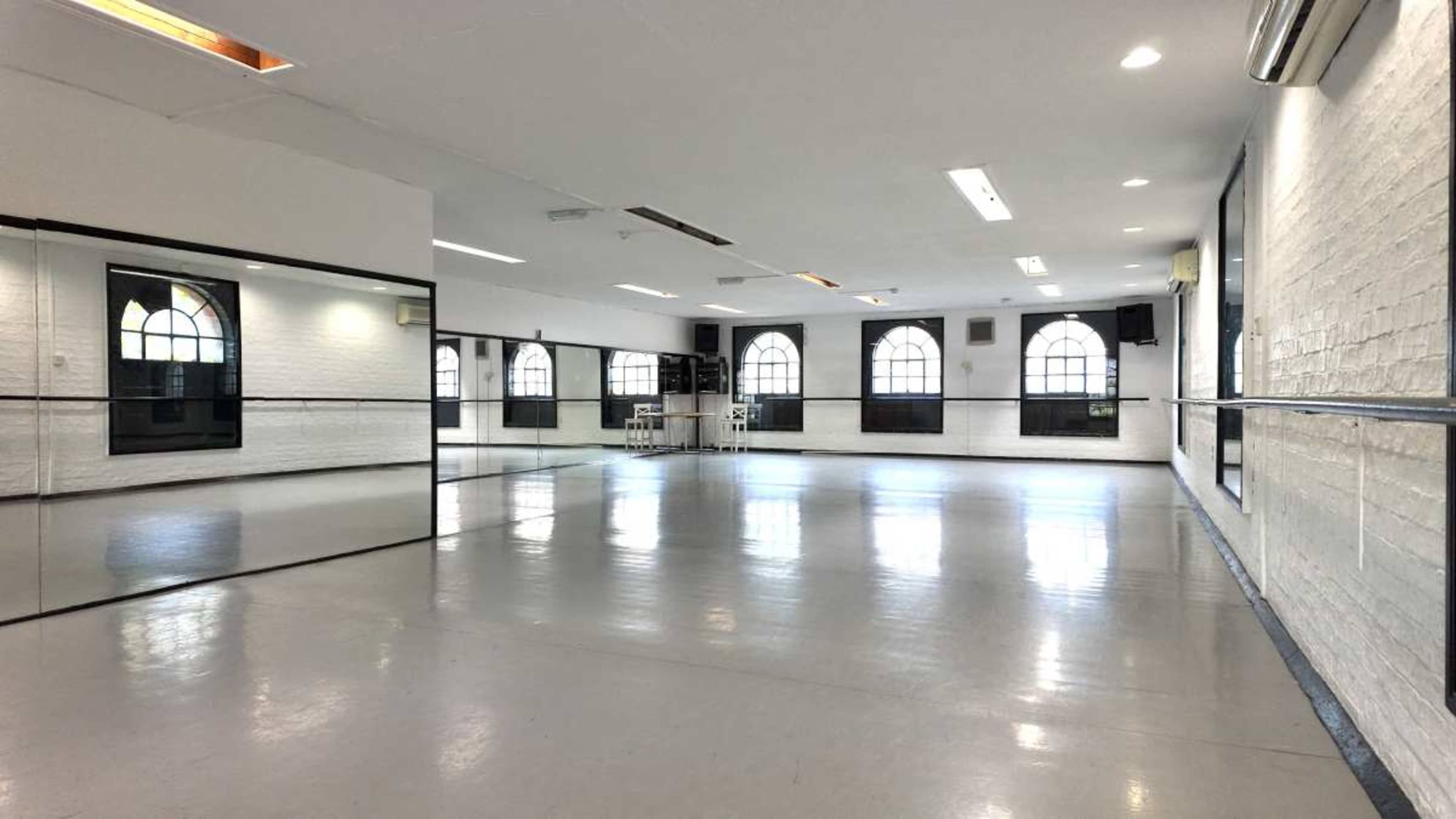 The image shows a spacious dance studio with large mirrors, white walls, and windows allowing natural light.