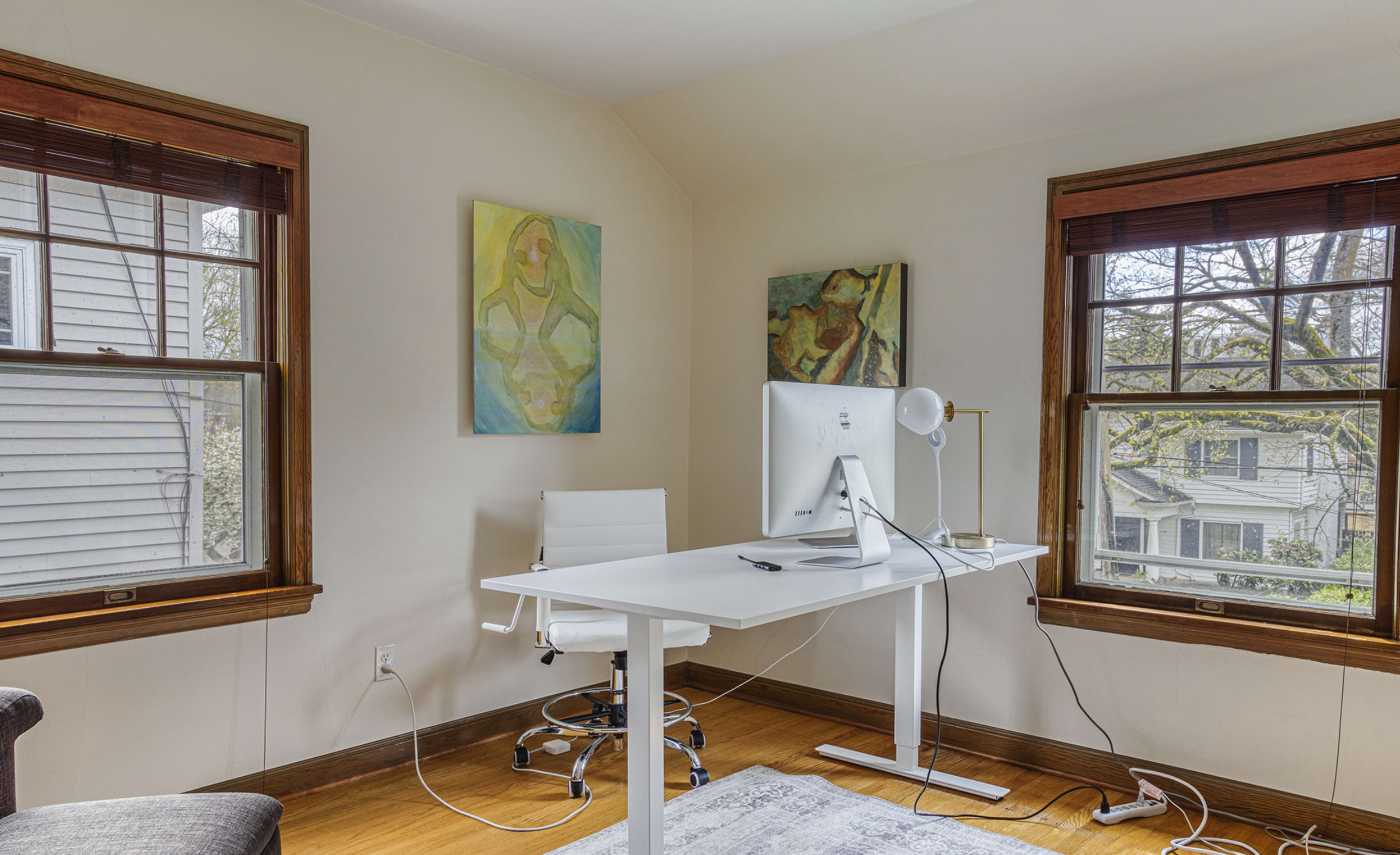 The image shows a modern home office with a white desk and an Apple computer situated between two framed artworks on the walls, illuminated by natural light from two windows.