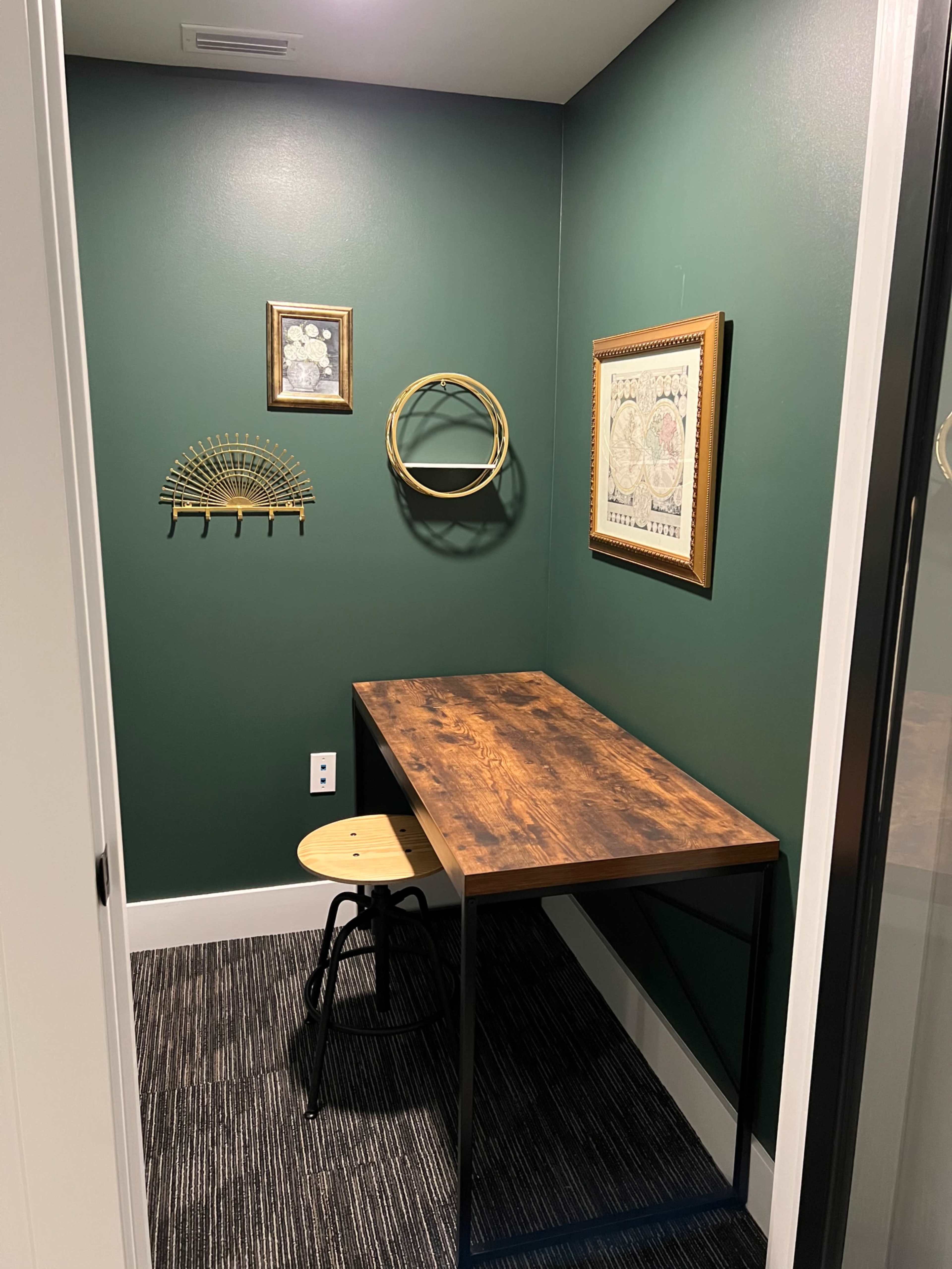 The image shows a small office space with a wooden desk and a stool, set against dark green walls adorned with framed wall art.