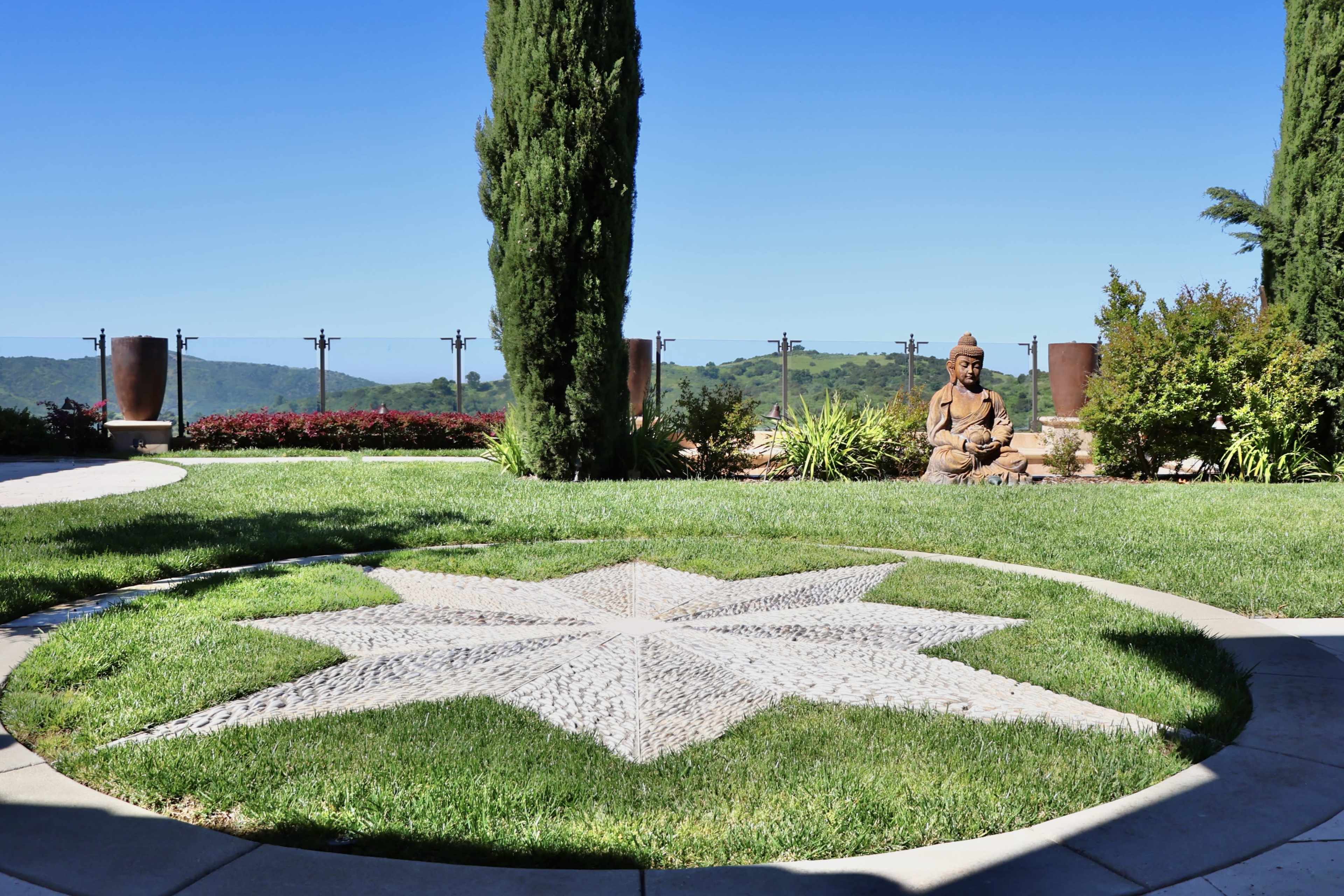 A landscaped garden featuring a stone star pattern embedded in grass, with a seated statue and lush greenery in the background.