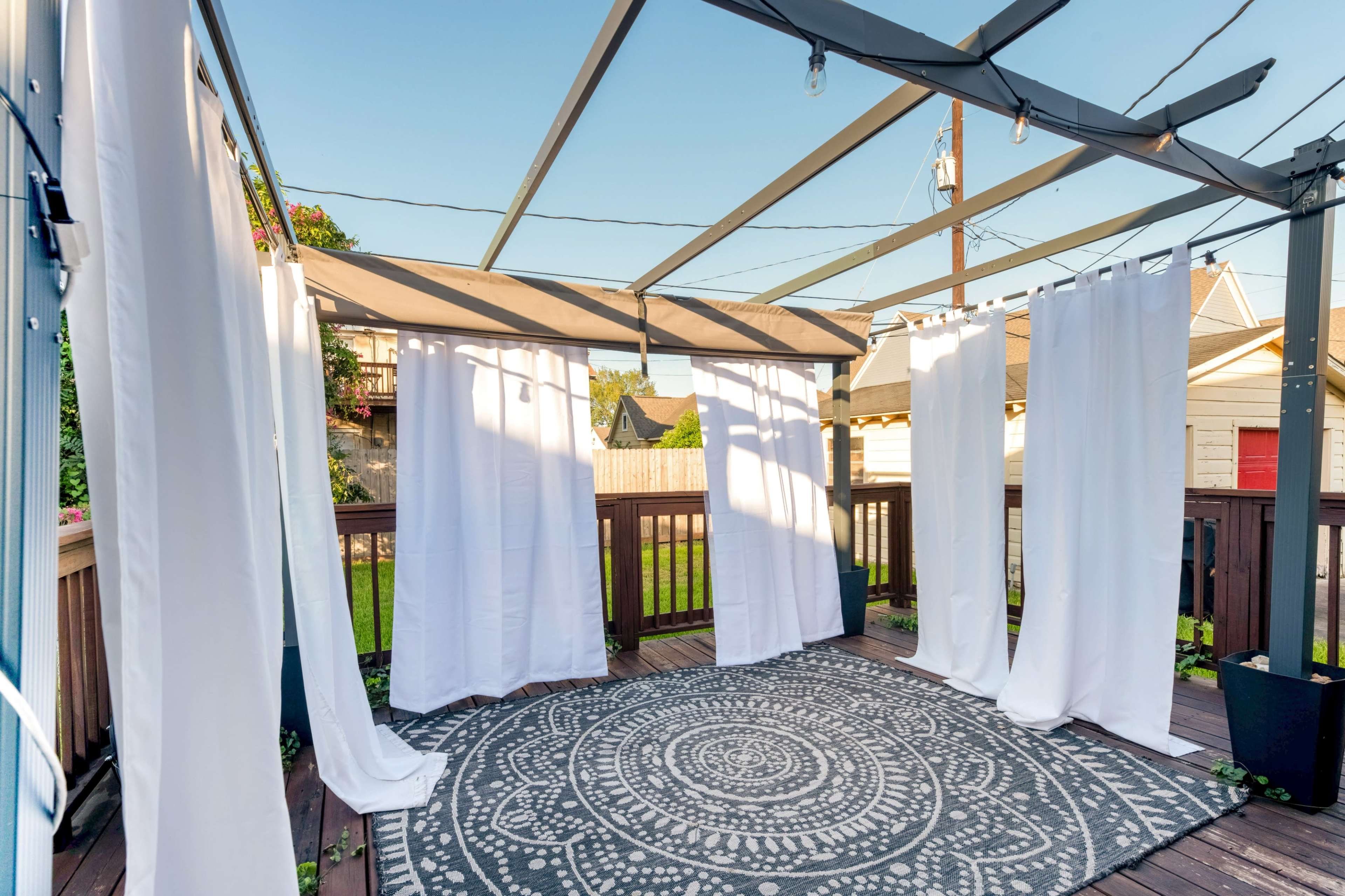 The image shows a covered wooden deck featuring white curtains hanging from a metal frame, with a round patterned rug on the floor.