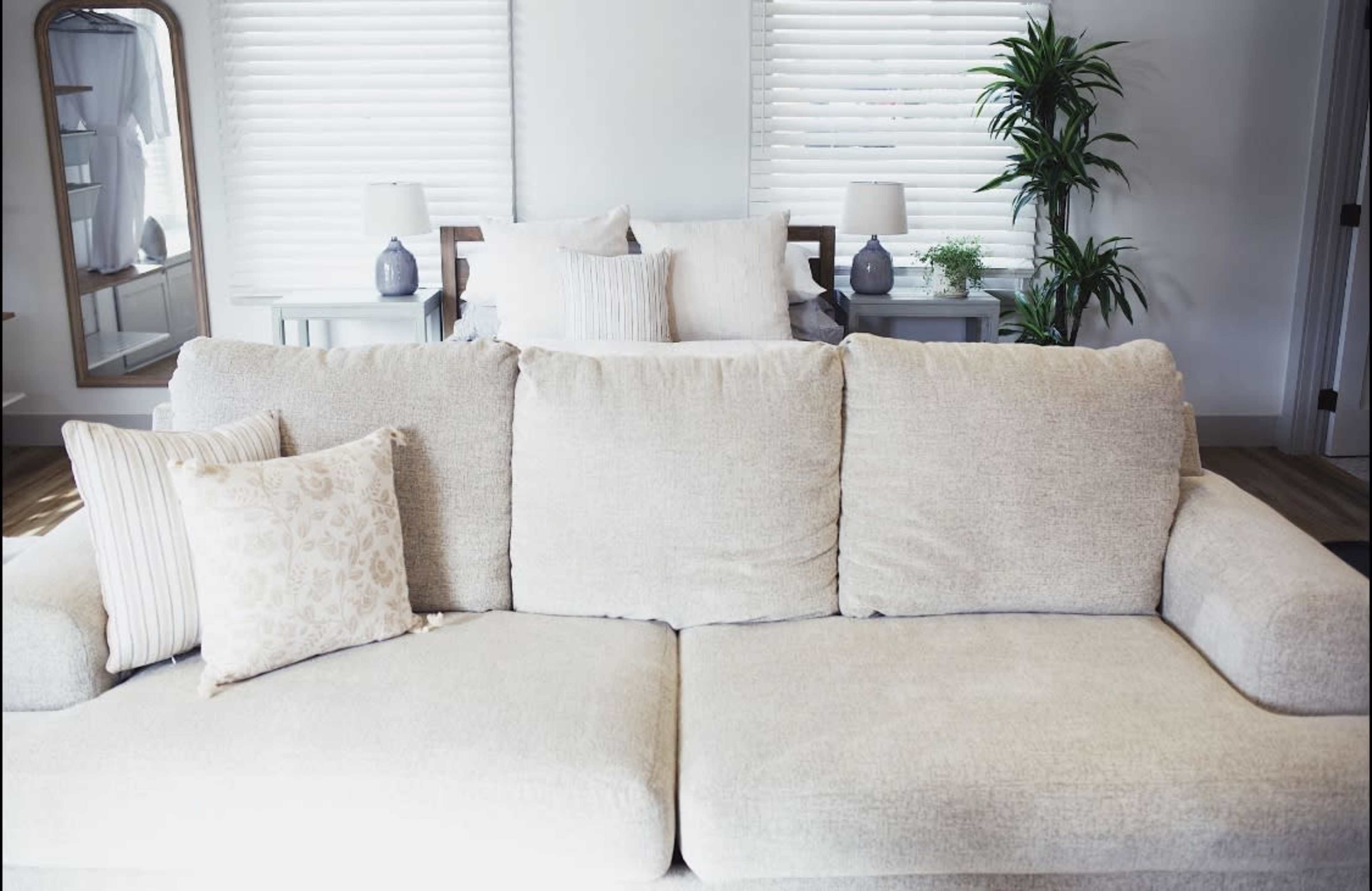 A light-colored sofa with decorative pillows is positioned in a bright room featuring two lamps and a potted plant.