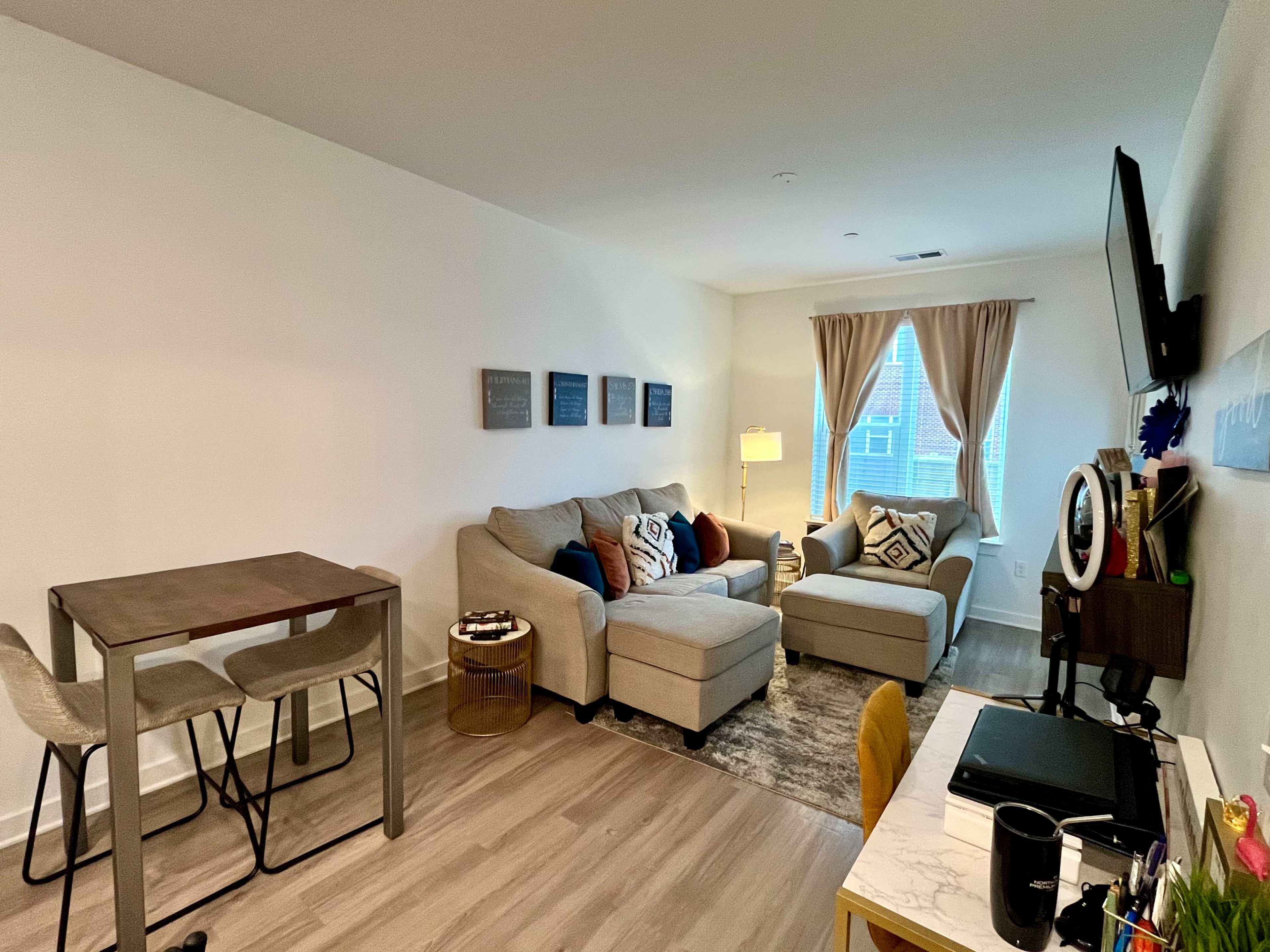 The image shows a modern living room setup with a beige sectional couch, a small dining table with two chairs, and a desk with a computer and a ring light.