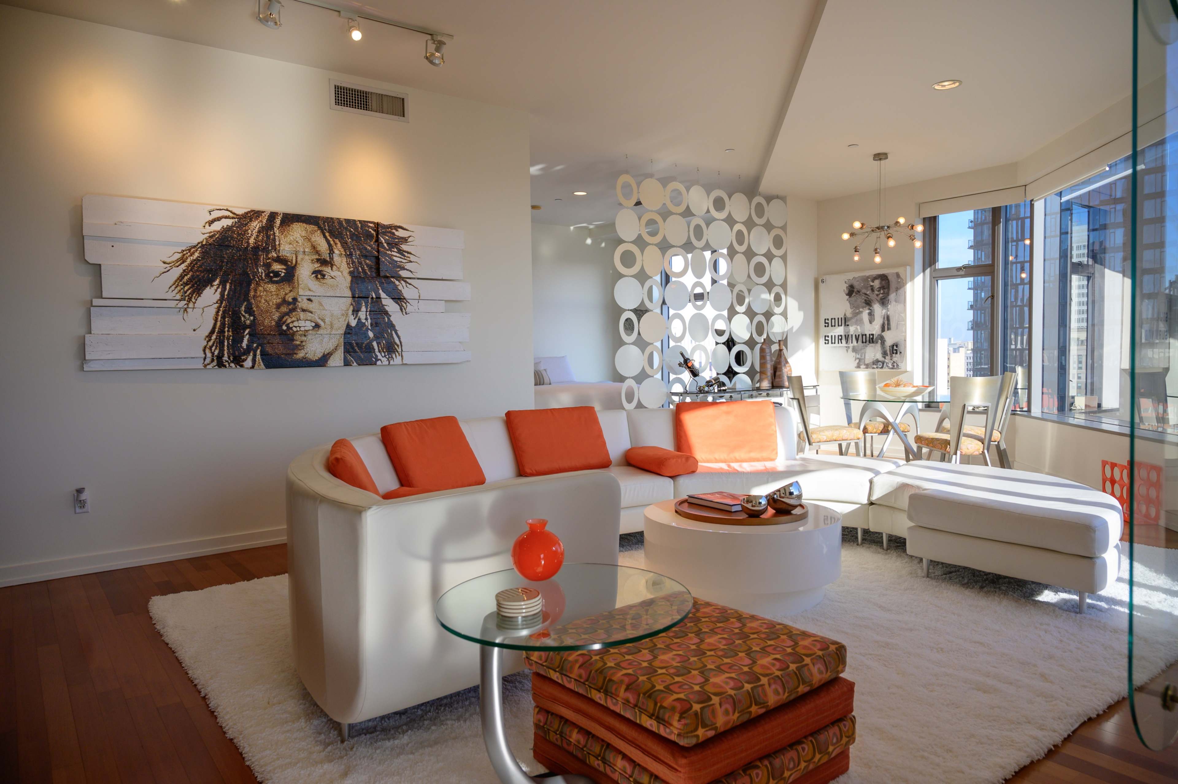 A modern living room features a white sectional sofa with orange cushions, a circular coffee table, and a large wall art piece, all complemented by large windows revealing a city view.