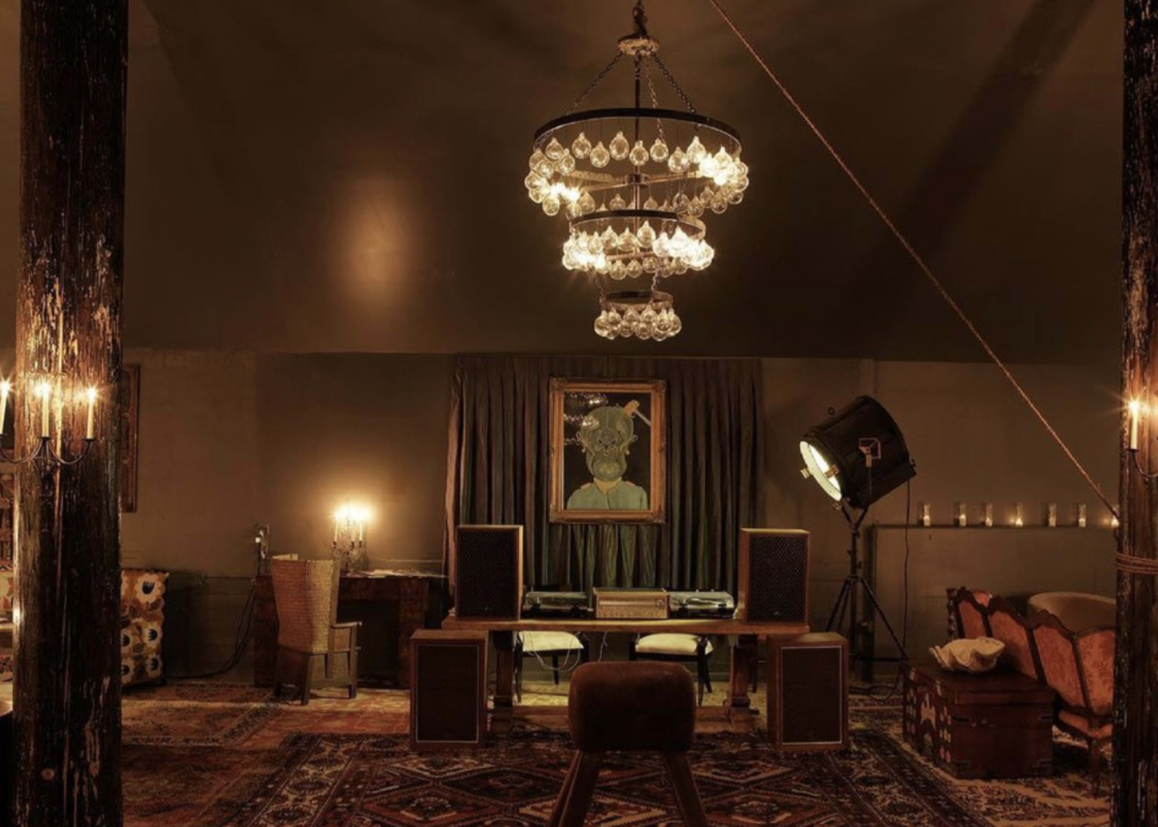 A dimly lit room features a large chandelier, a painting on the wall, a wooden desk with speakers, and various seating areas.