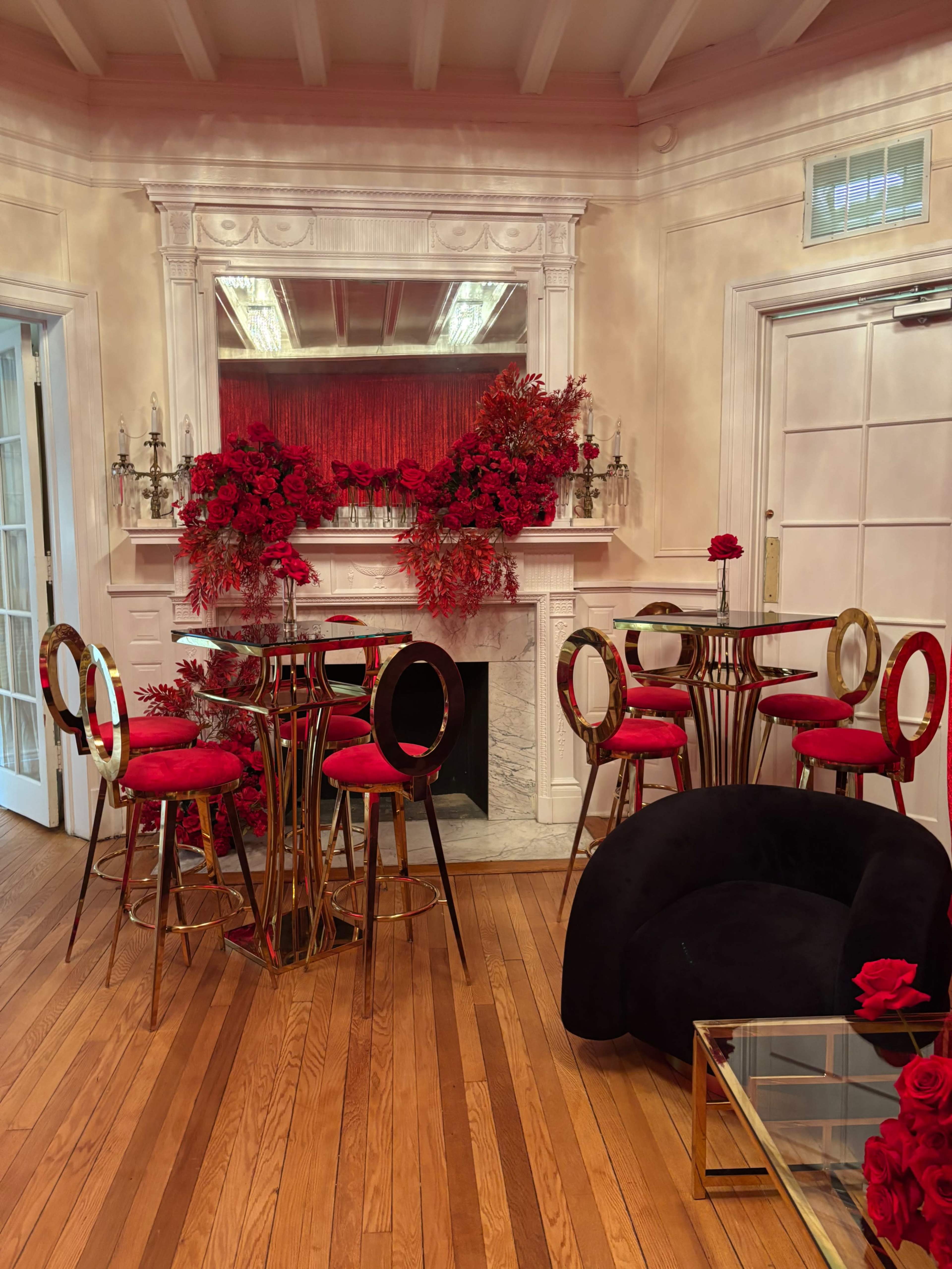 The room features a cozy seating area with a black chair and a set of red bar stools arranged around a gold and glass table, adorned with decorative red flowers around a fireplace.
