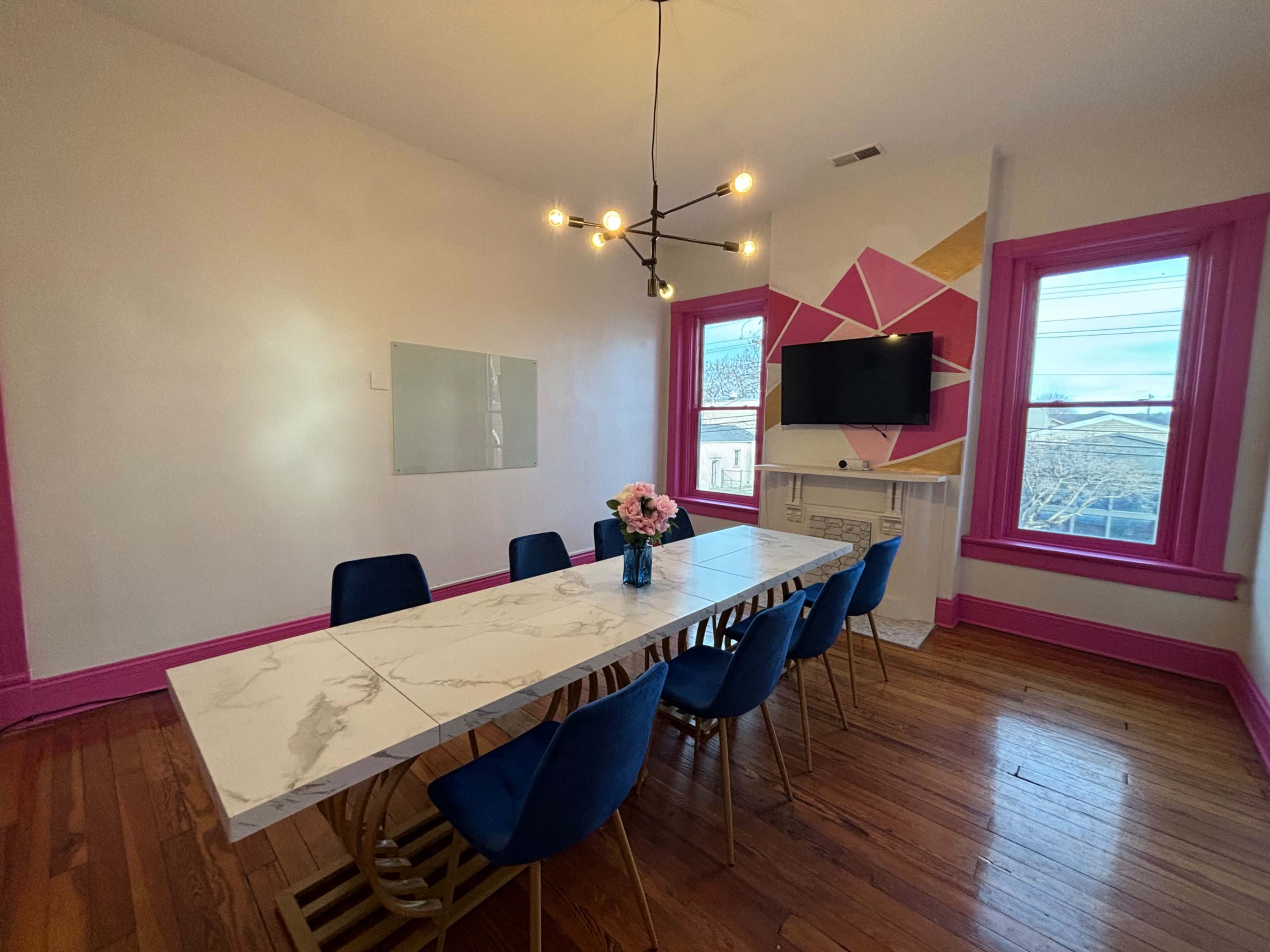 A dining room with a long marble table surrounded by blue chairs, a floral centerpiece, and pink-accented walls featuring a television and a geometric design.