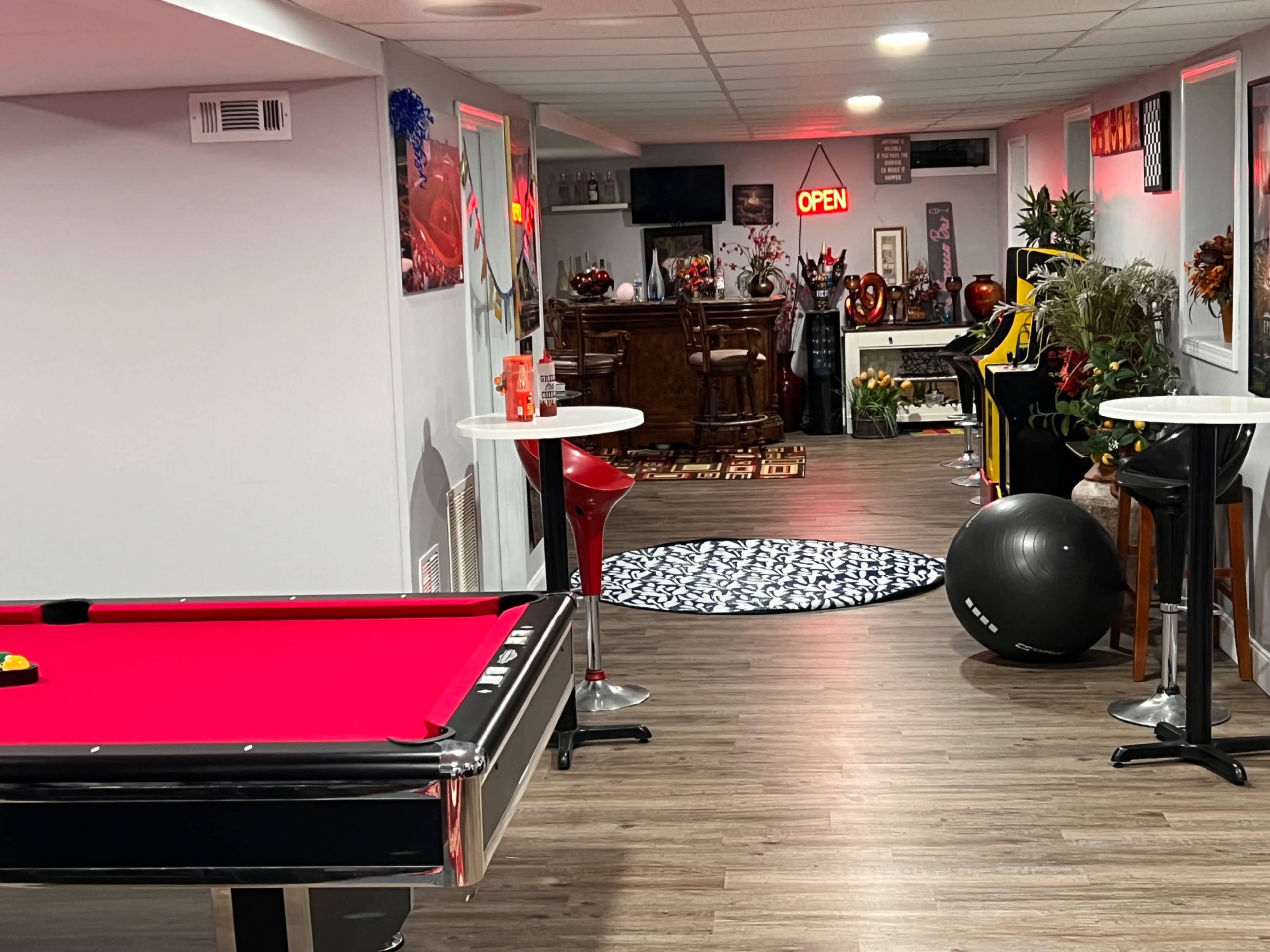 The image shows a basement game room featuring a red pool table, a bar area, and various decorative elements, with dim lighting and a large rug on the floor.