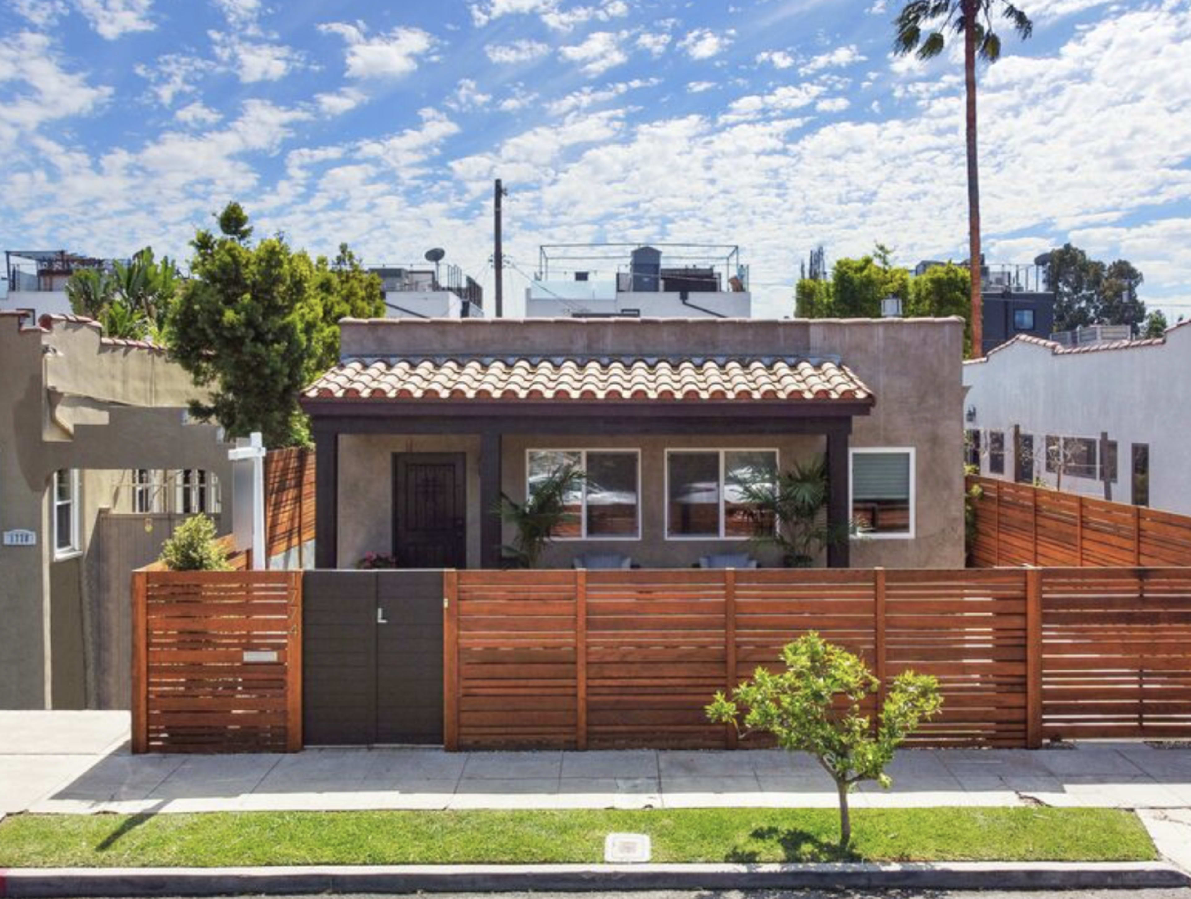 A single-story house with a tiled roof and a wooden fence, surrounded by palm trees and partly cloudy skies.