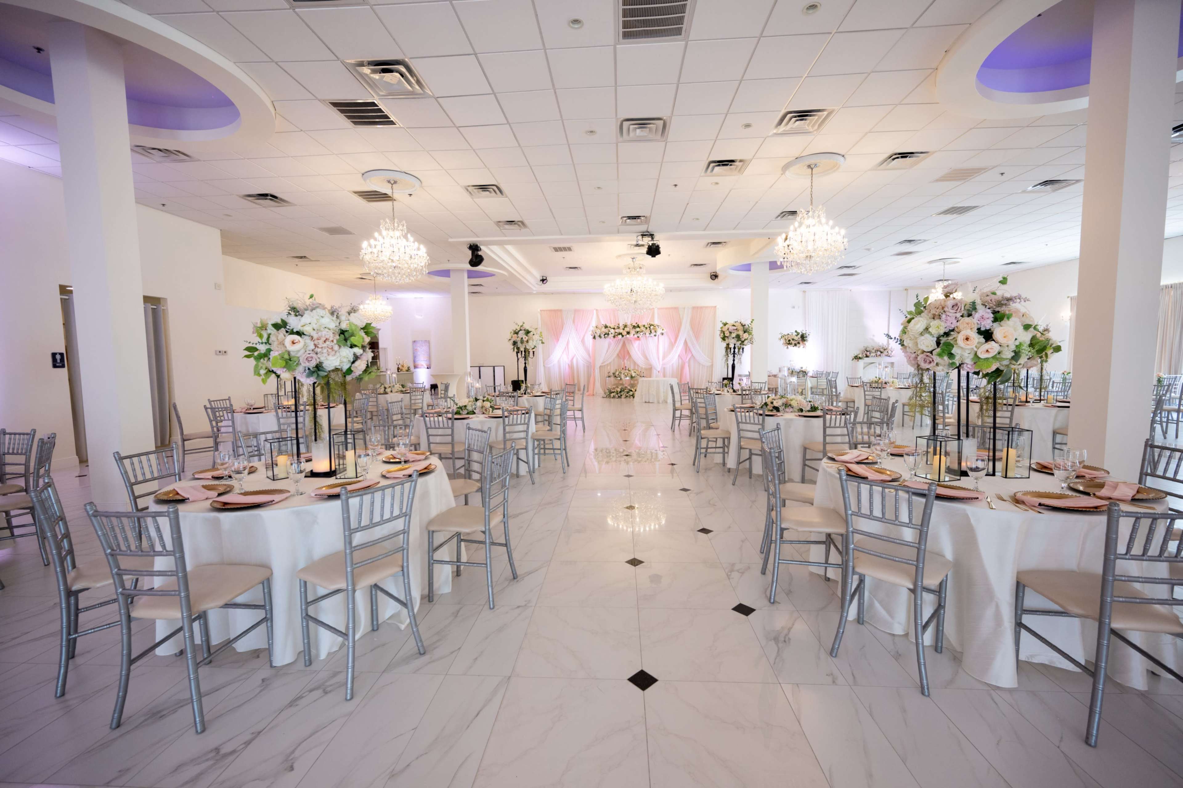 The image depicts a spacious, elegantly decorated event hall with round tables set for a formal gathering, adorned with floral centerpieces and chandeliers overhead.