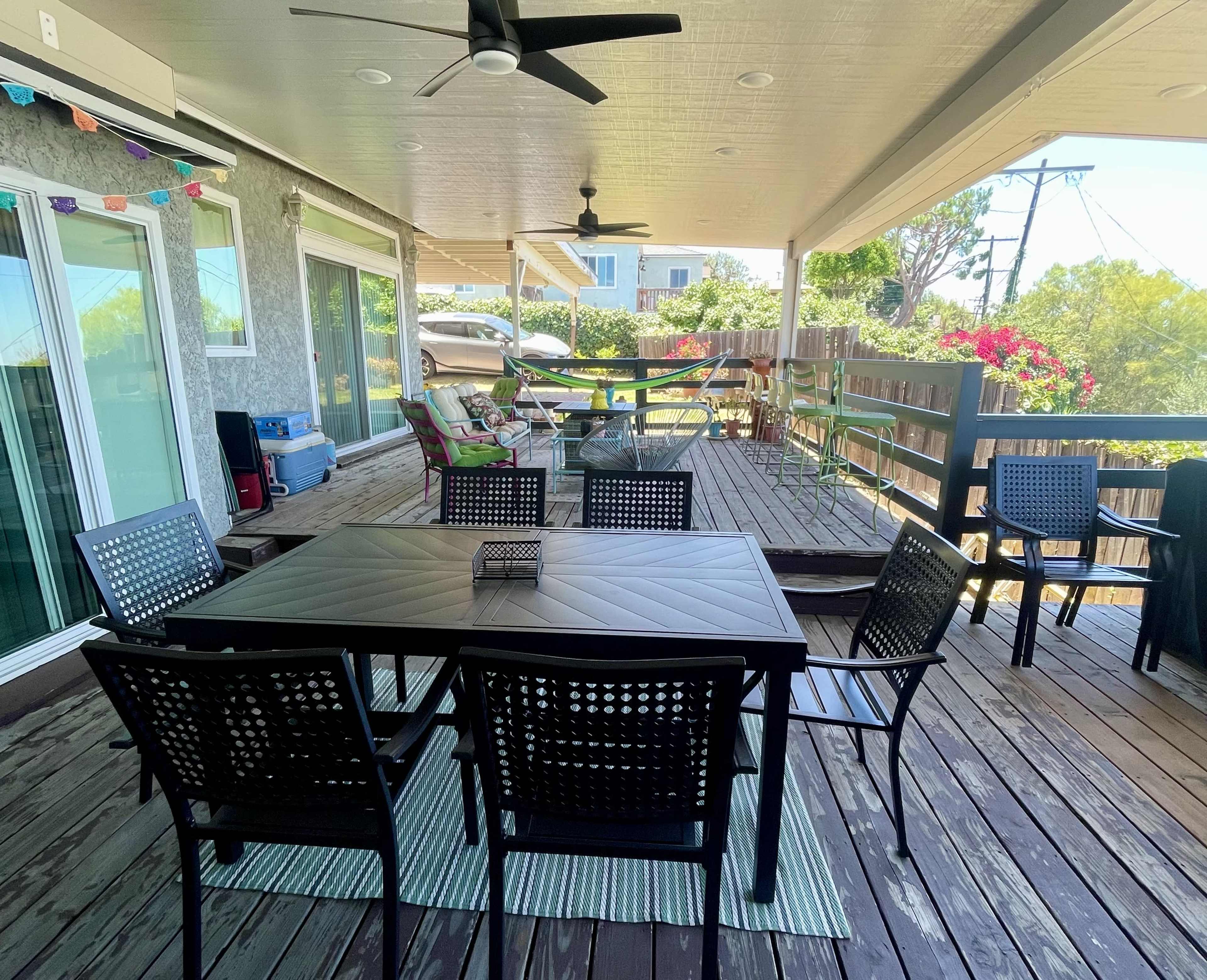 The image shows a spacious outdoor patio with a dining table and chairs, surrounded by greenery and colorful decorations.