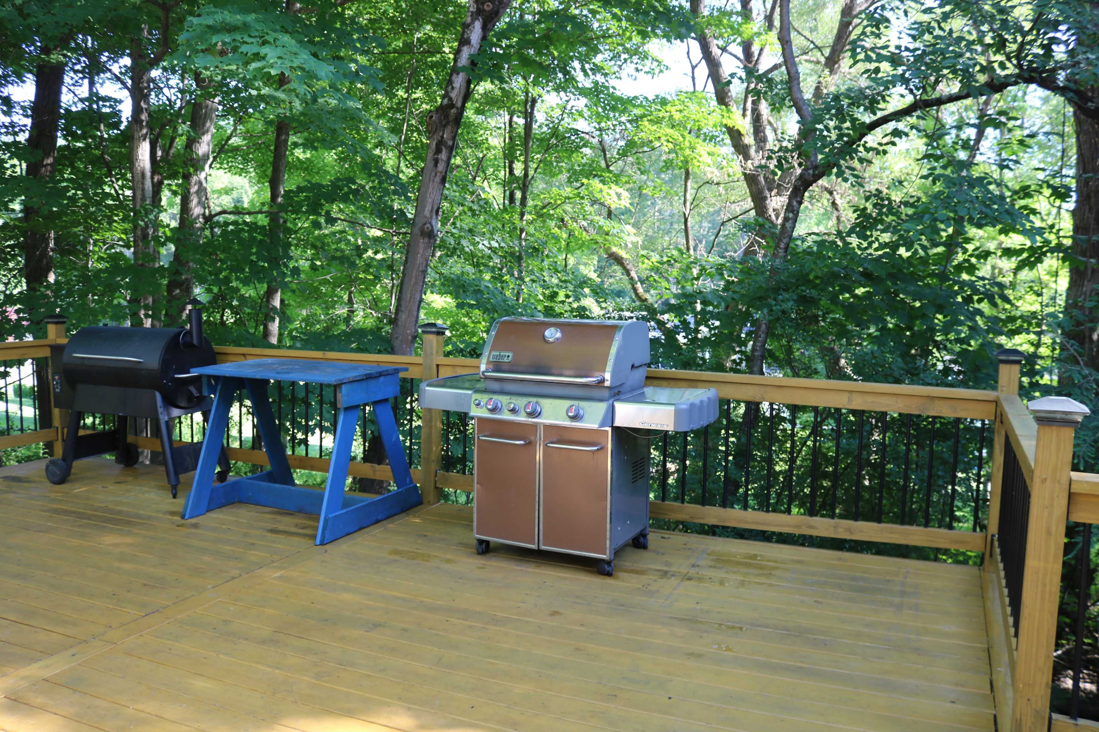 A wooden deck with a blue work table and a stainless steel grill, surrounded by lush green trees.