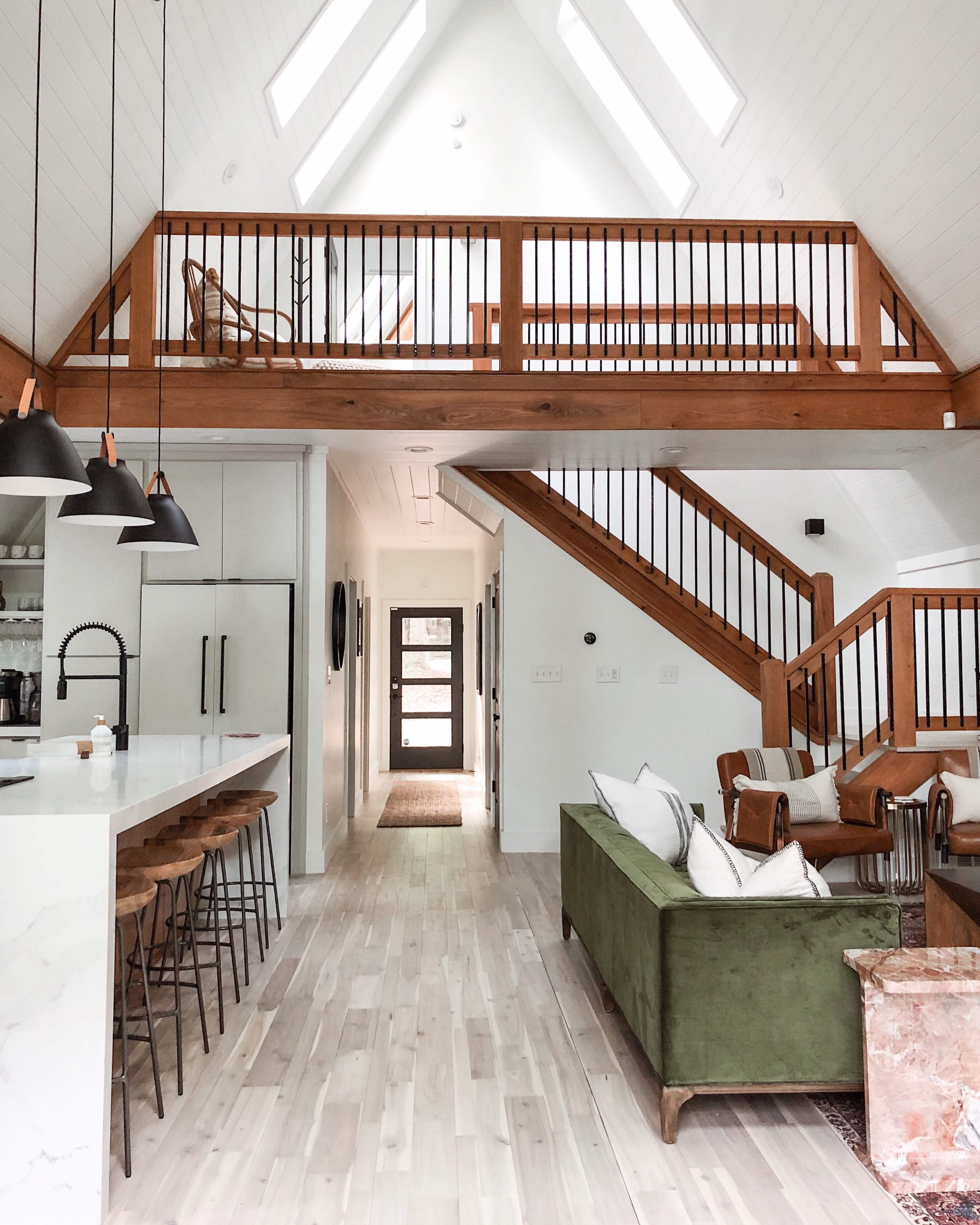 A modern interior of a light-filled home with a two-story layout, featuring a kitchen bar with seating, a staircase, and an open living area with a green couch.