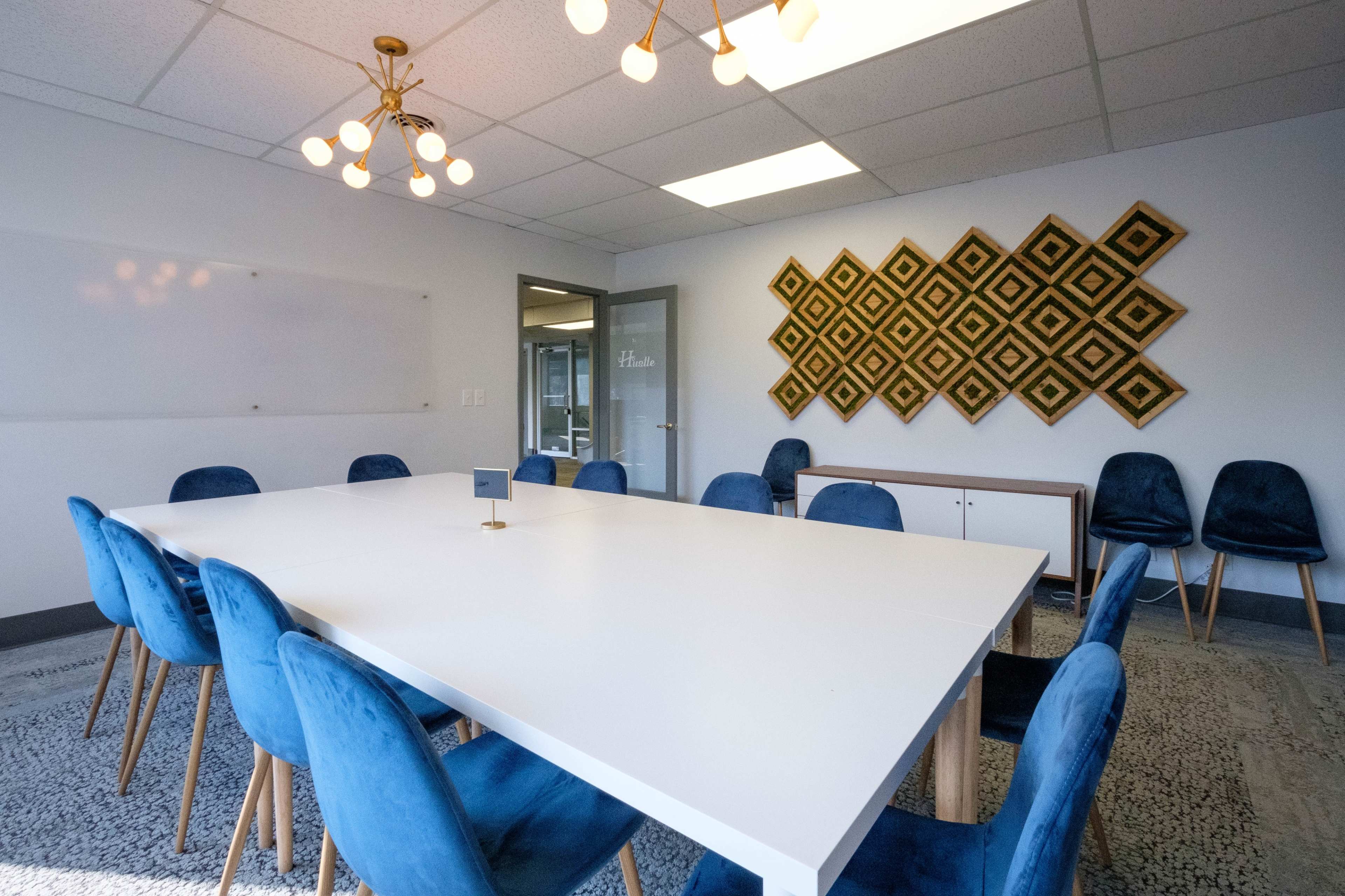 A spacious conference room features a large white table surrounded by blue chairs, with a decorative wall hanging in the background.