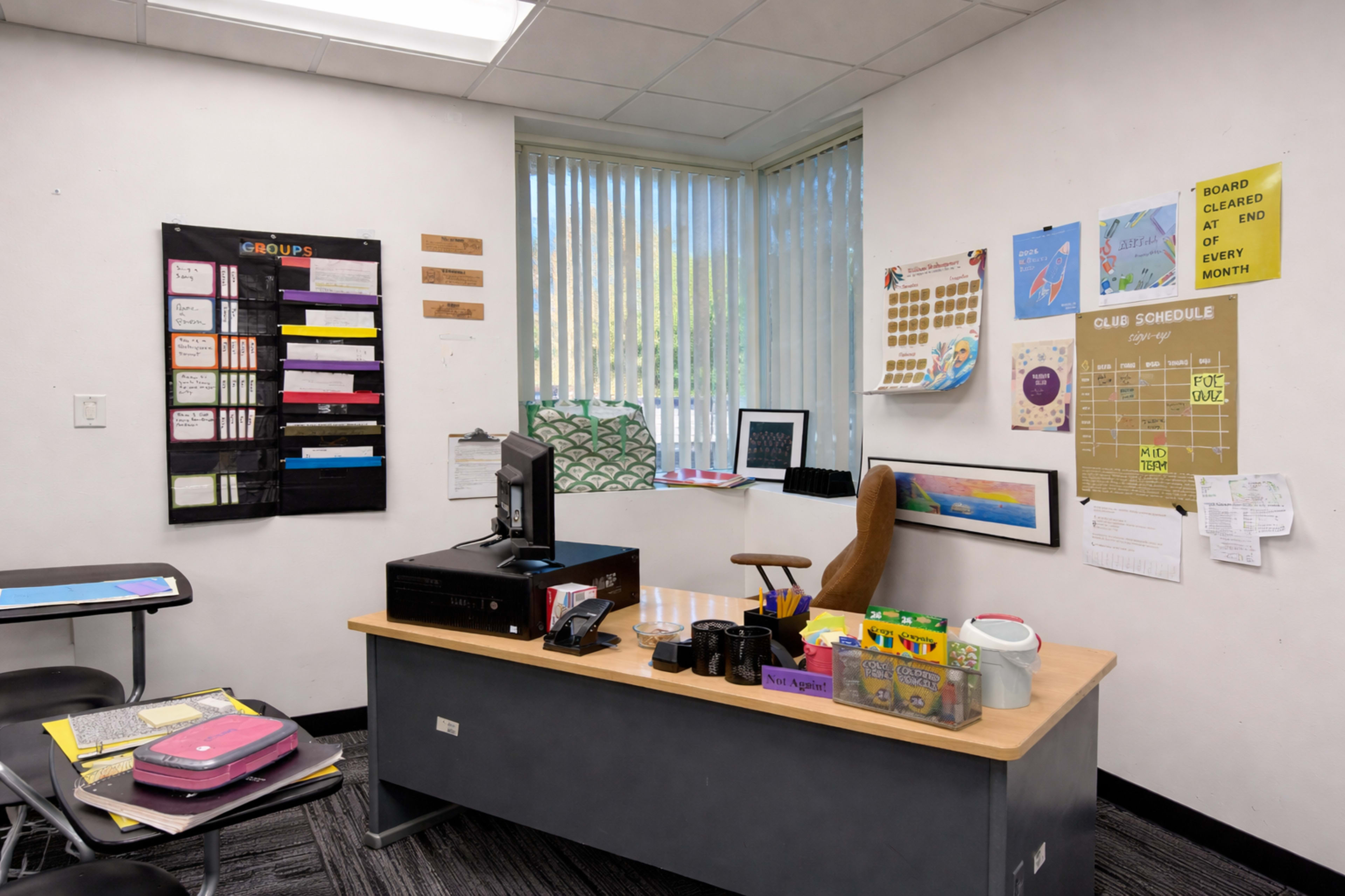 A classroom features a desk with a computer, various organizational materials on the walls, and a window allowing natural light.
