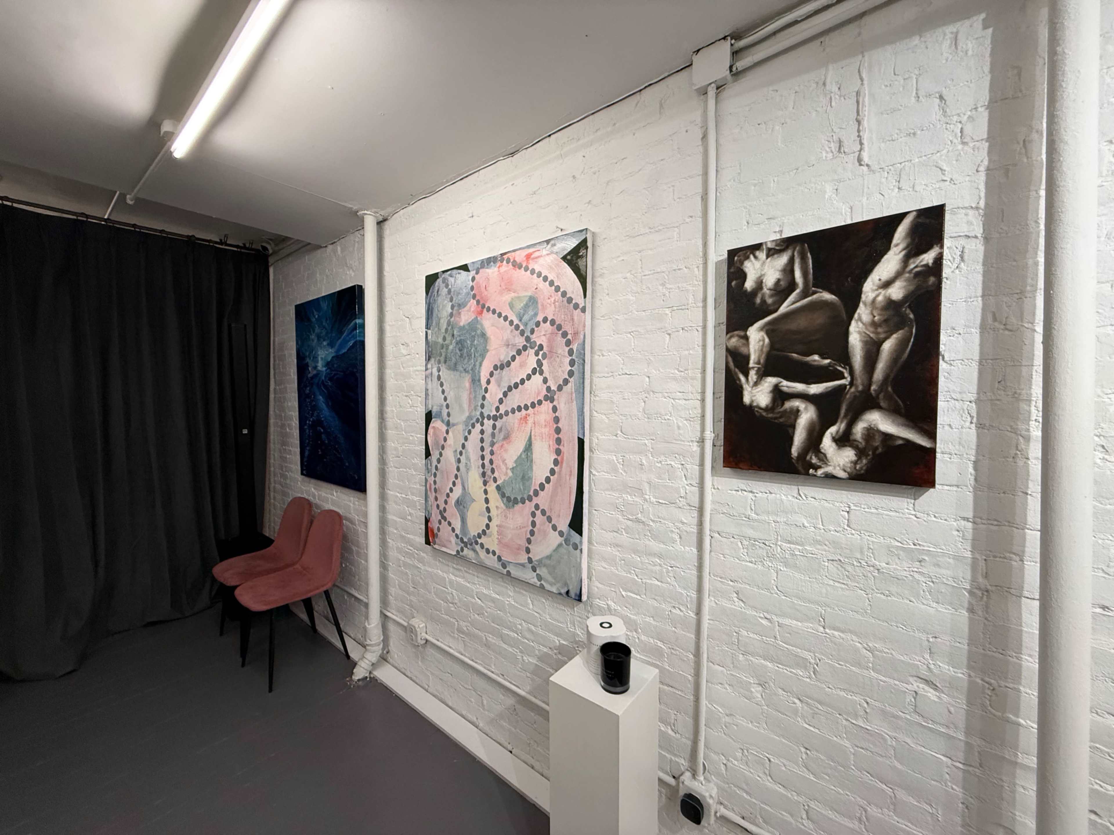 The image shows an art gallery interior featuring four pieces of artwork displayed on a white brick wall, along with two pink chairs and a black curtain.