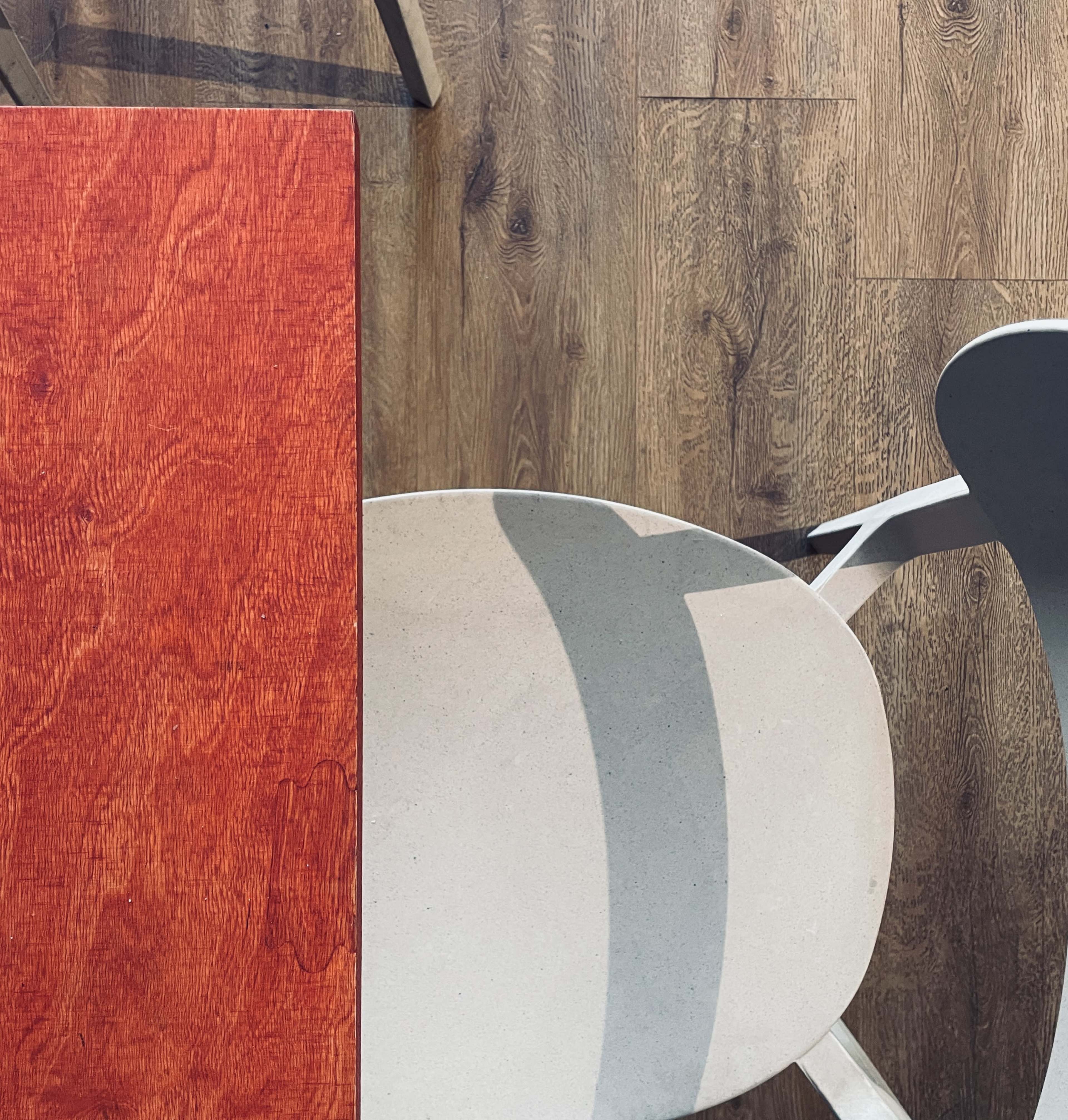 A wooden table with a red surface is positioned above a light-colored chair on a wooden floor.