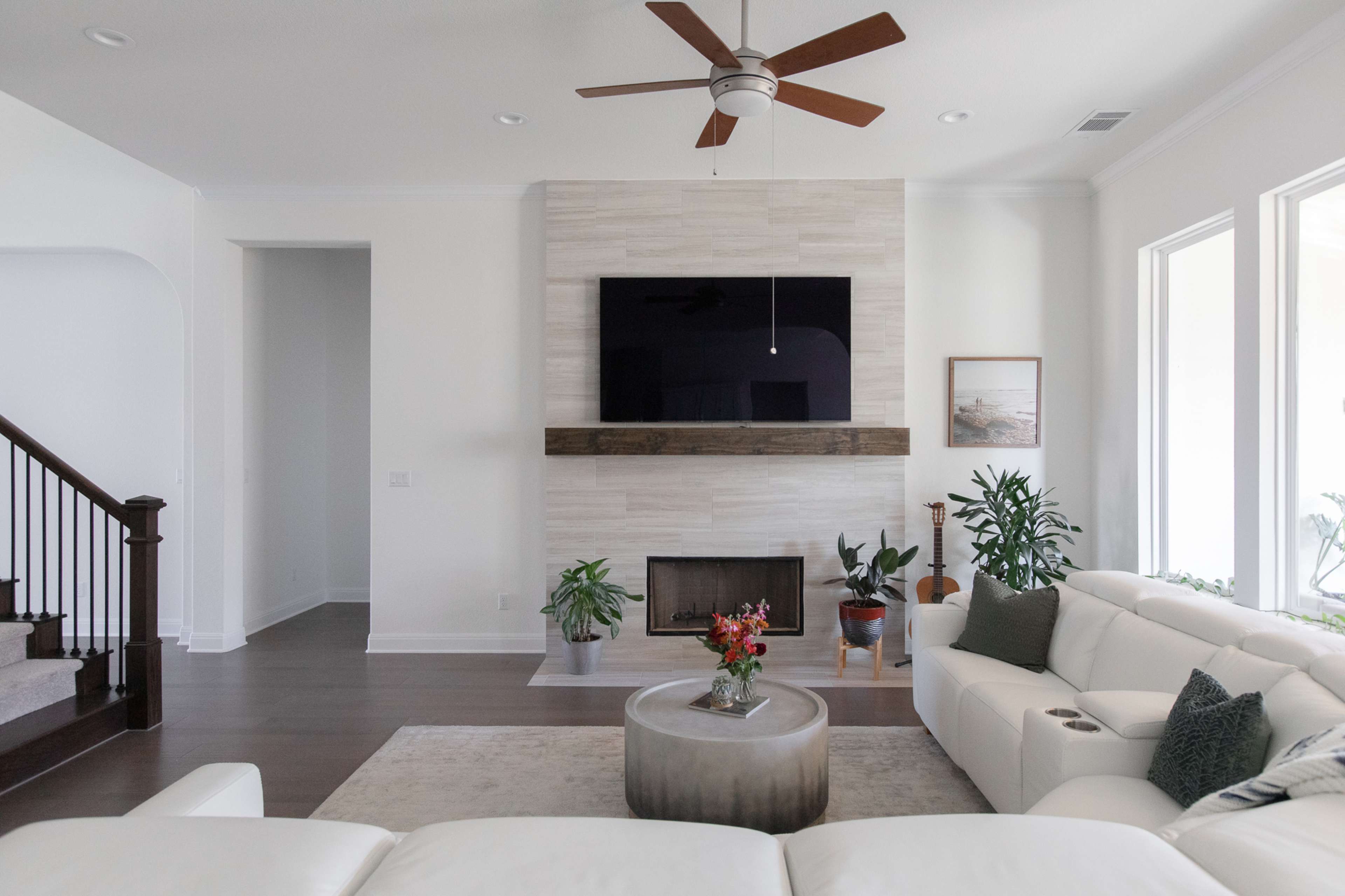 A modern living room featuring a white sectional sofa, a circular coffee table, a wall-mounted television, and a fireplace with plants and decorative items.