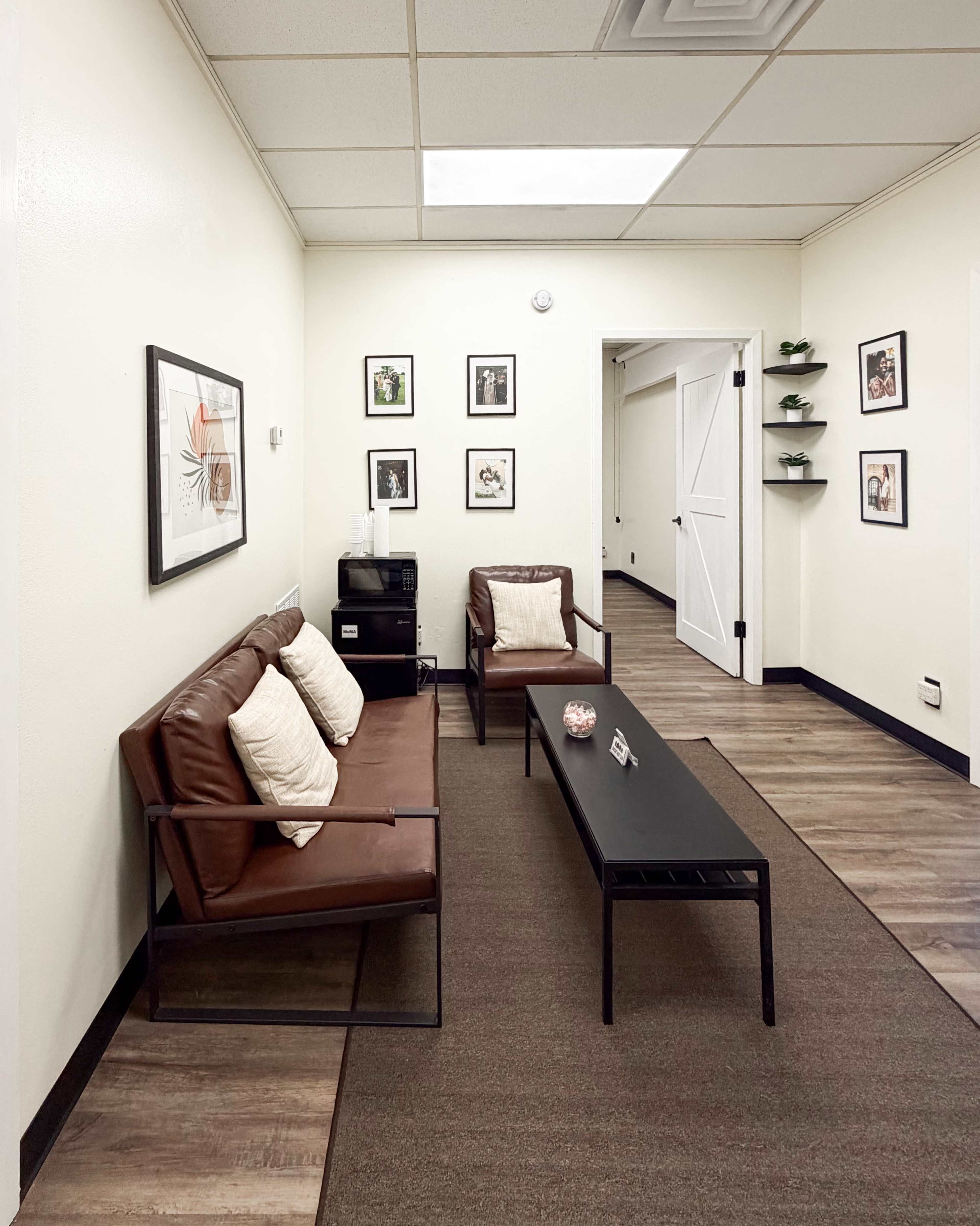 The image shows a small waiting area with two brown chairs, a black coffee table, framed pictures on the walls, and a door at the end of the corridor.