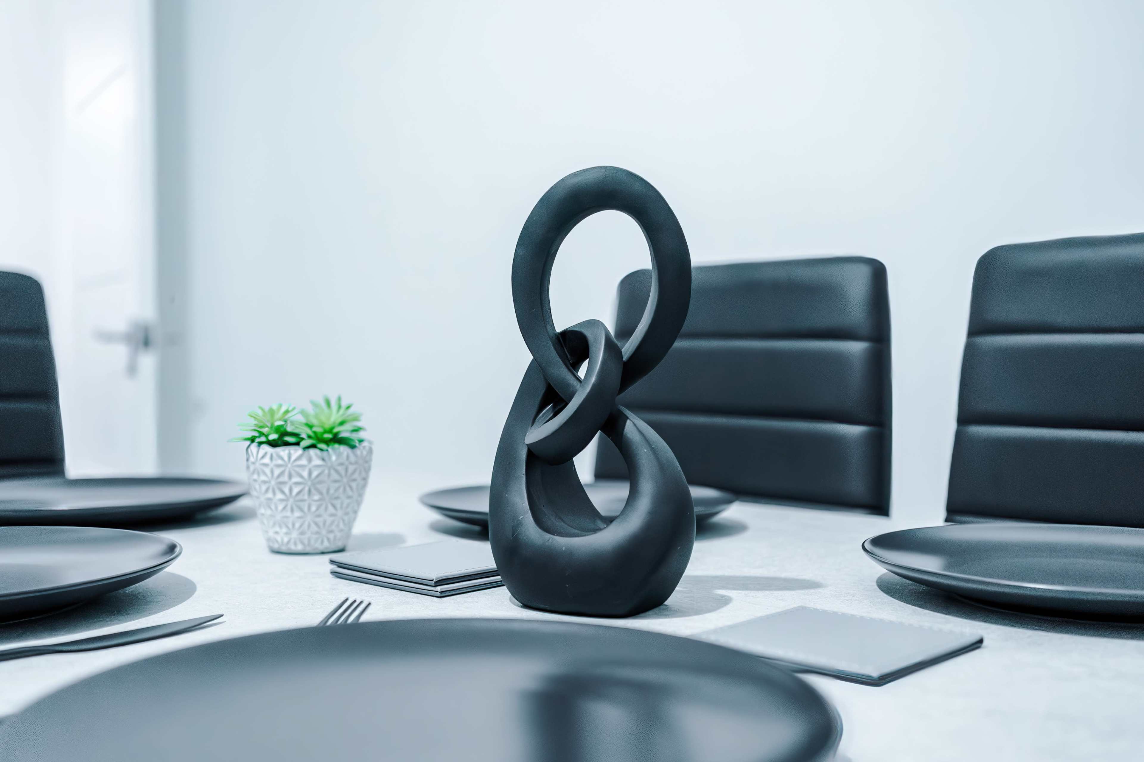 A modern dining table is set with black plates and four upholstered chairs, featuring a sculptural centerpiece and a small potted plant.