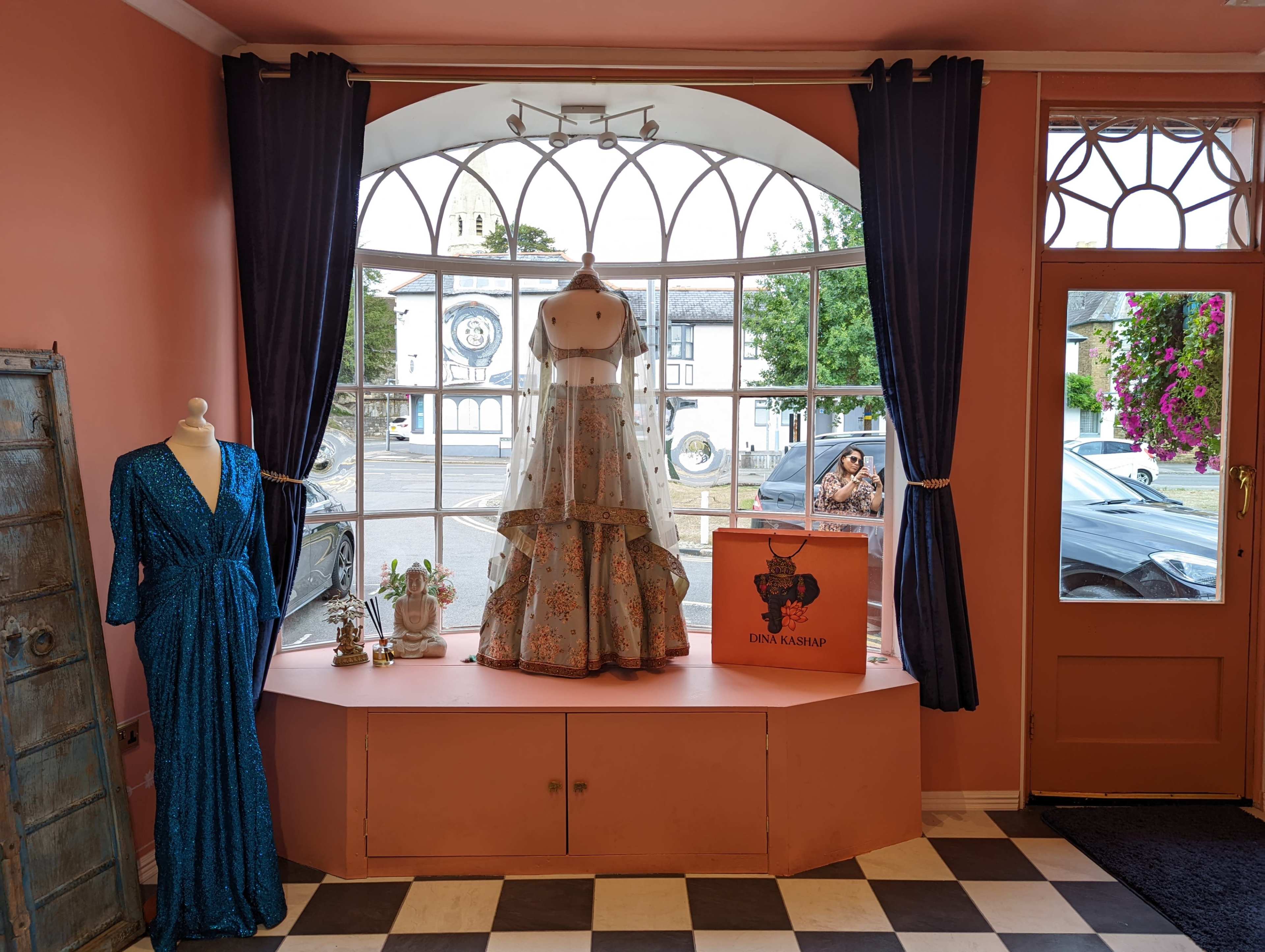 A boutique window display featuring a long dress on a mannequin and a blue dress hanging nearby, with large windows providing a view of the outside.