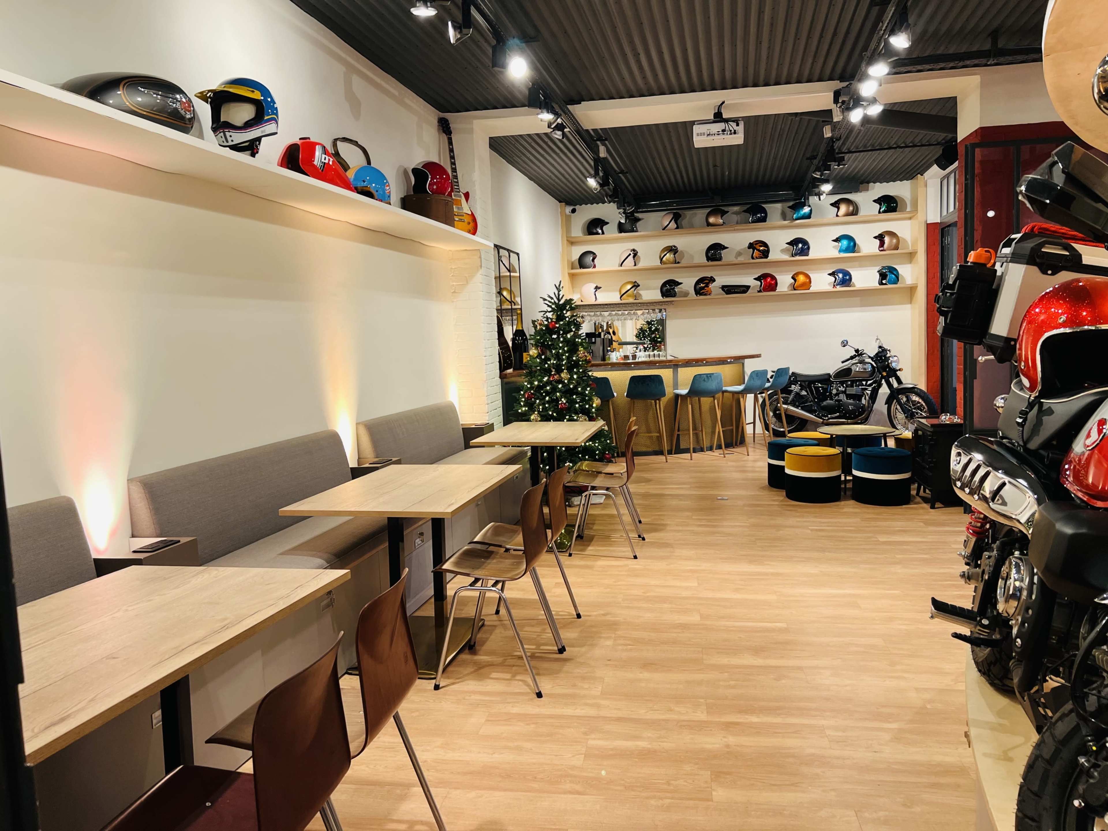 The image shows a modern cafe or store interior with wooden tables and chairs, a decorative Christmas tree, shelves displaying various motorcycle helmets, and a motorcycle in the background.