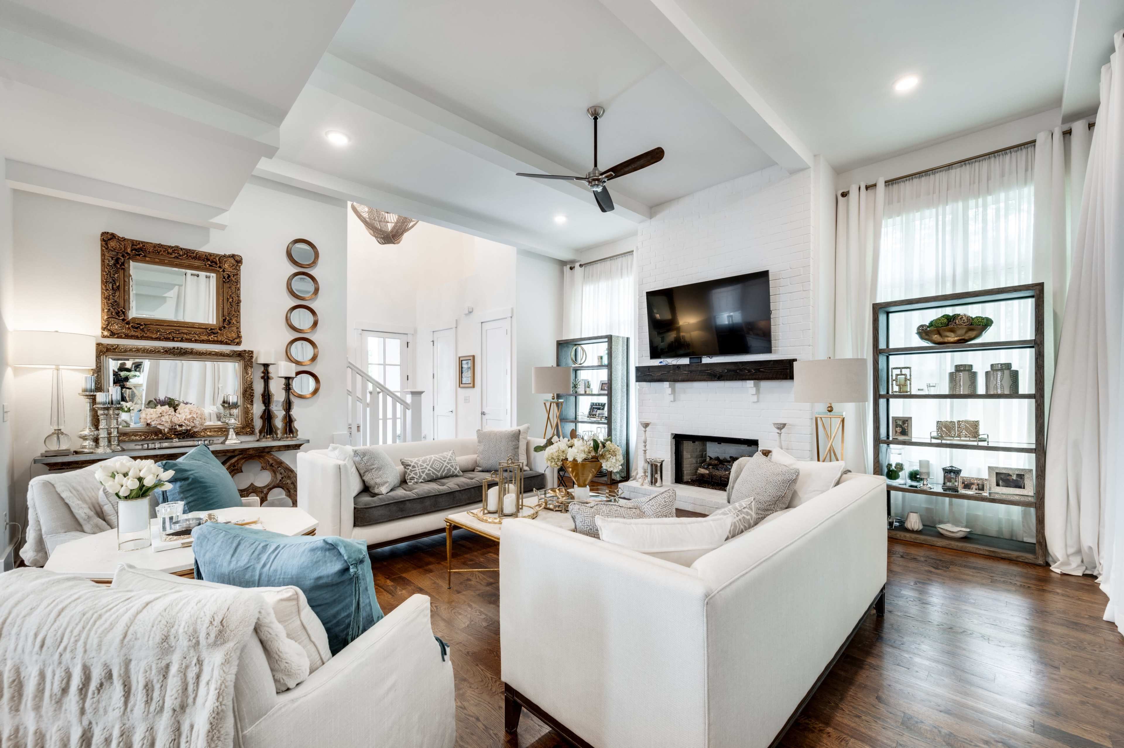 The image shows a bright and modern living room with light-colored sofas, a television mounted on the wall, and decorative shelves displaying various items.