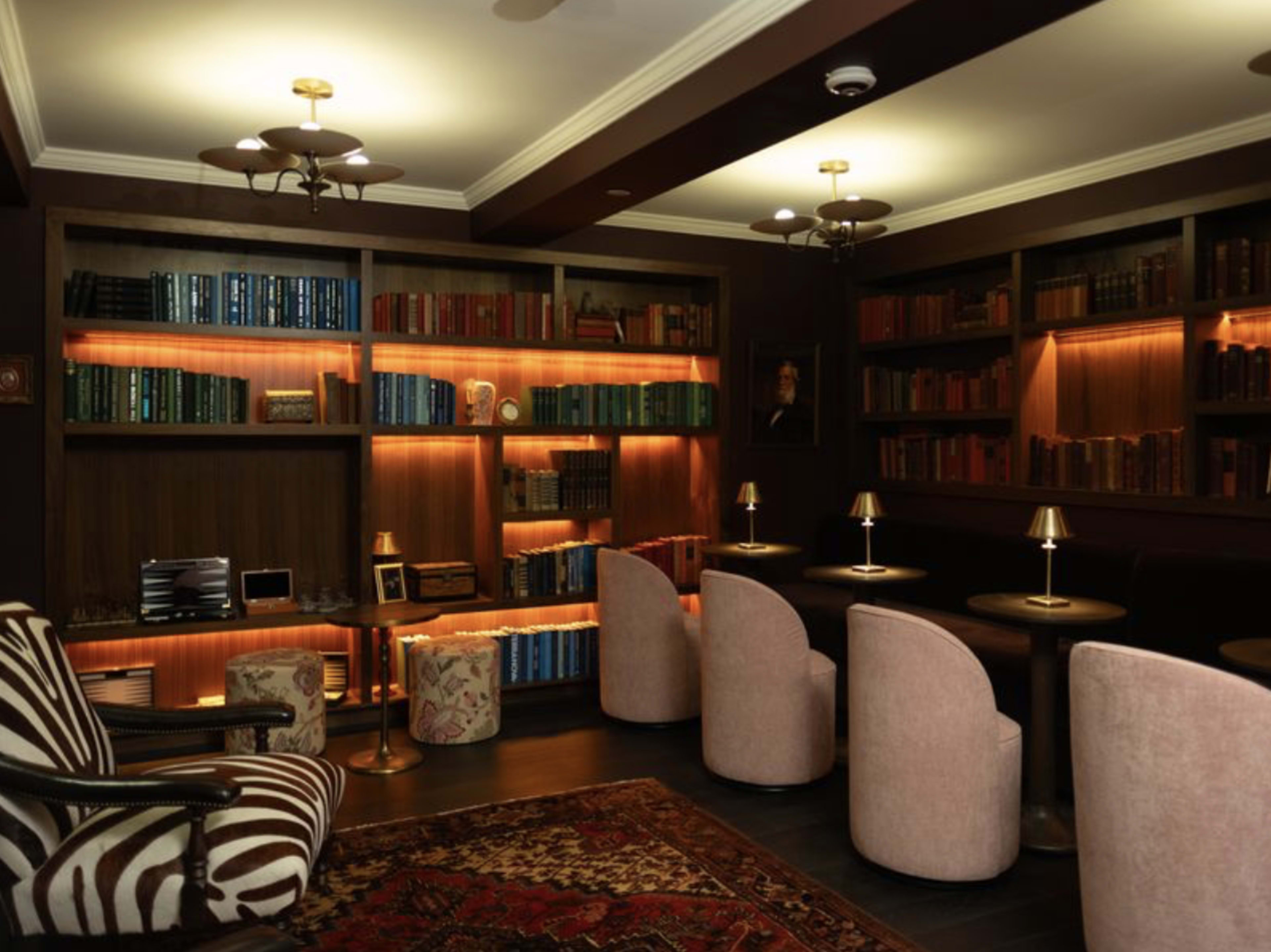 The image shows a dimly lit library with wooden shelves filled with books, plush seating, and stylish lamps.