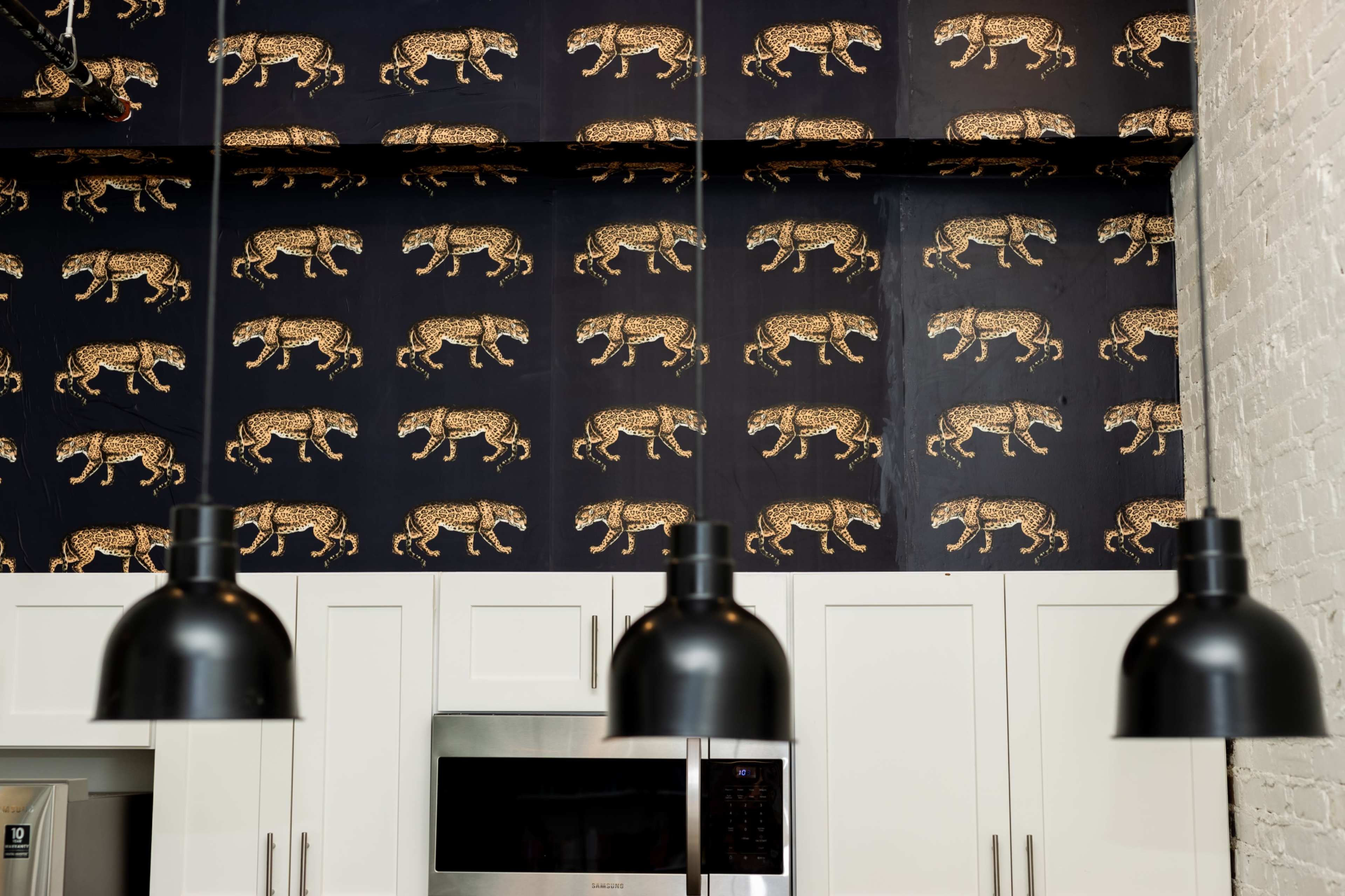 A kitchen with a patterned wallpaper depicting leopards and black pendant lights hanging above white cabinetry.