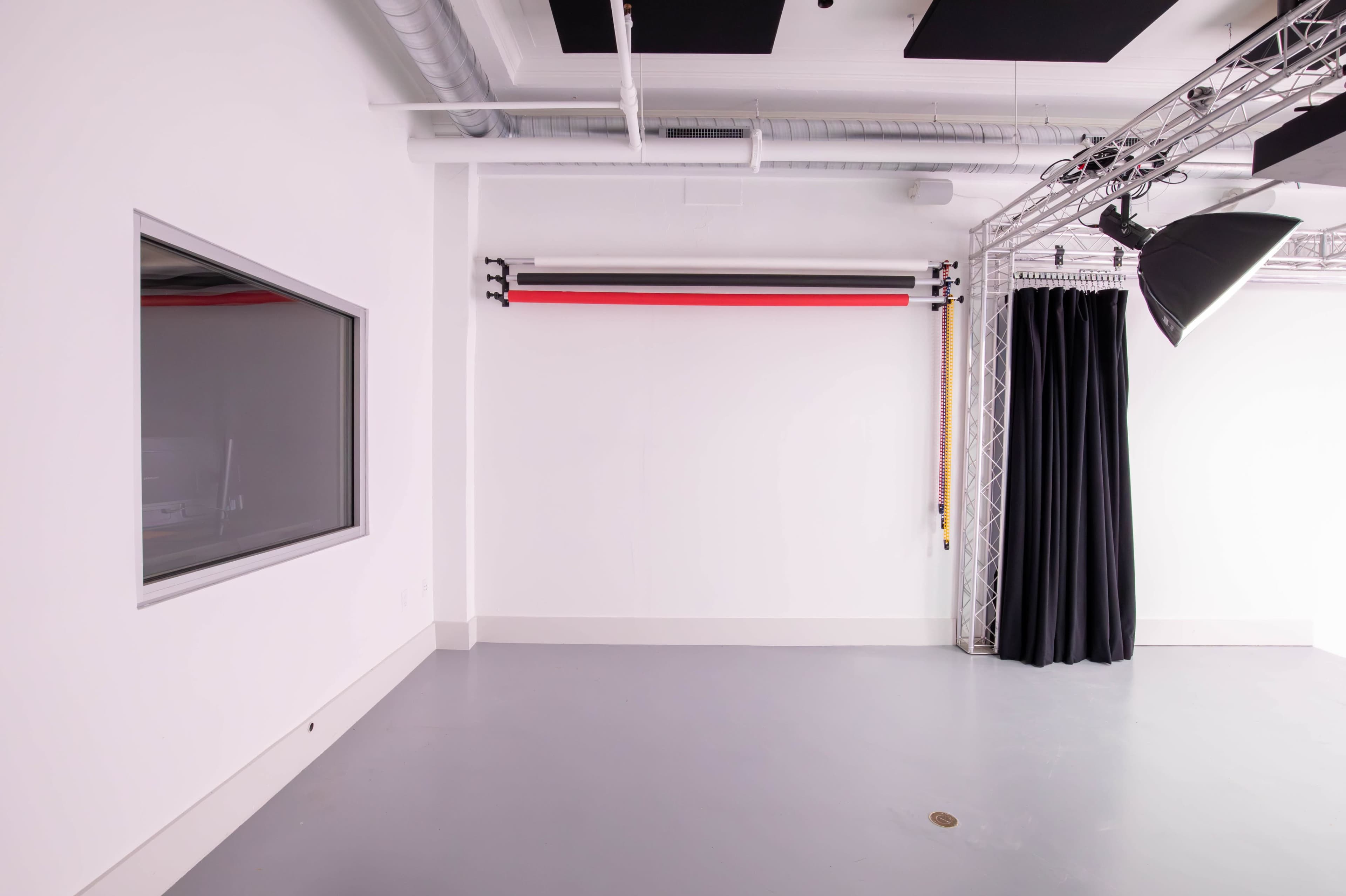 The image shows a spacious, minimalist studio with a gray floor, a large window, a curtain, and a backdrop support system mounted on the wall.