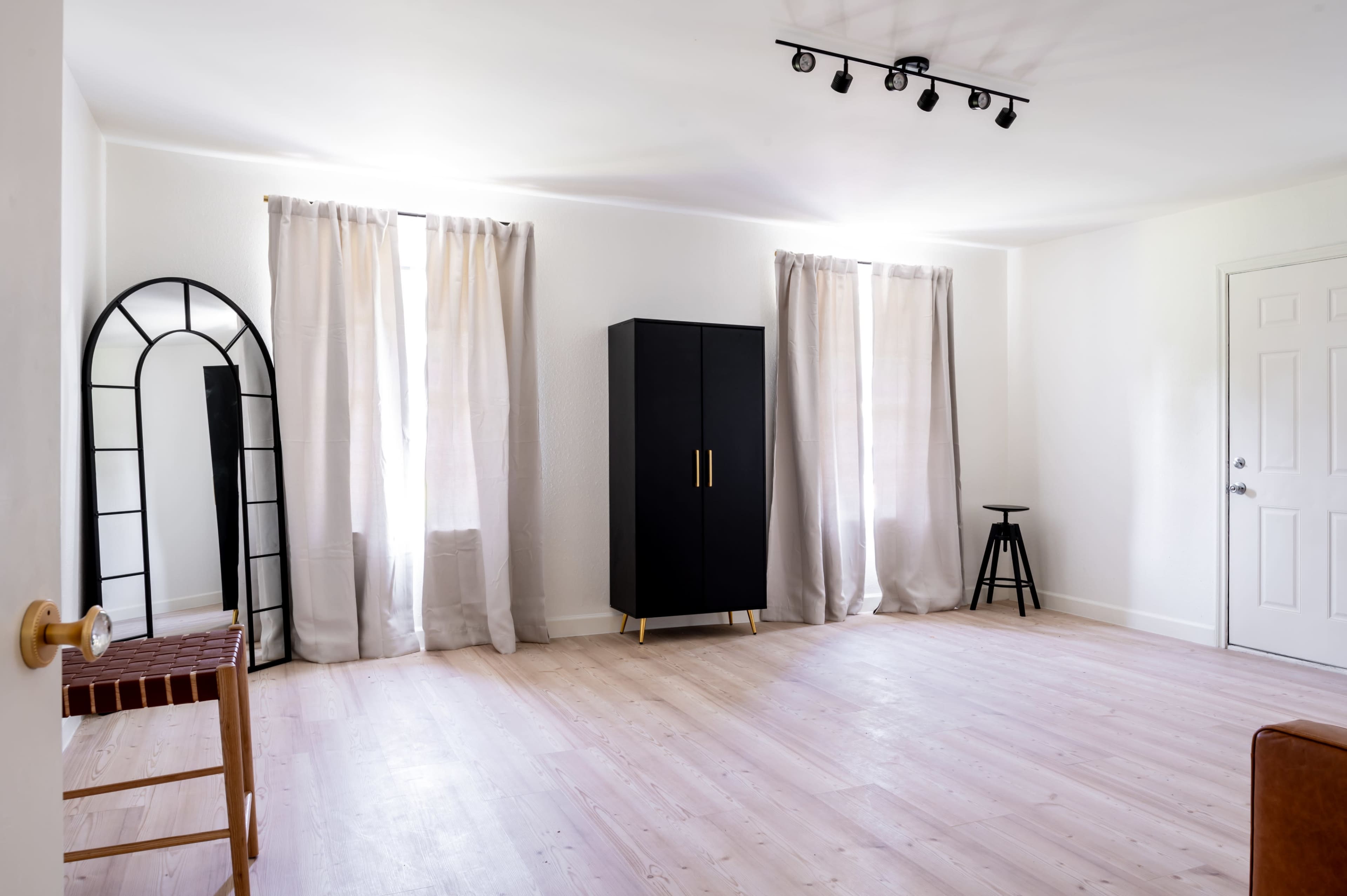 The room features two windows with curtains, a tall black cabinet, a stool, a full-length mirror, and a wooden chair on a light-colored floor.