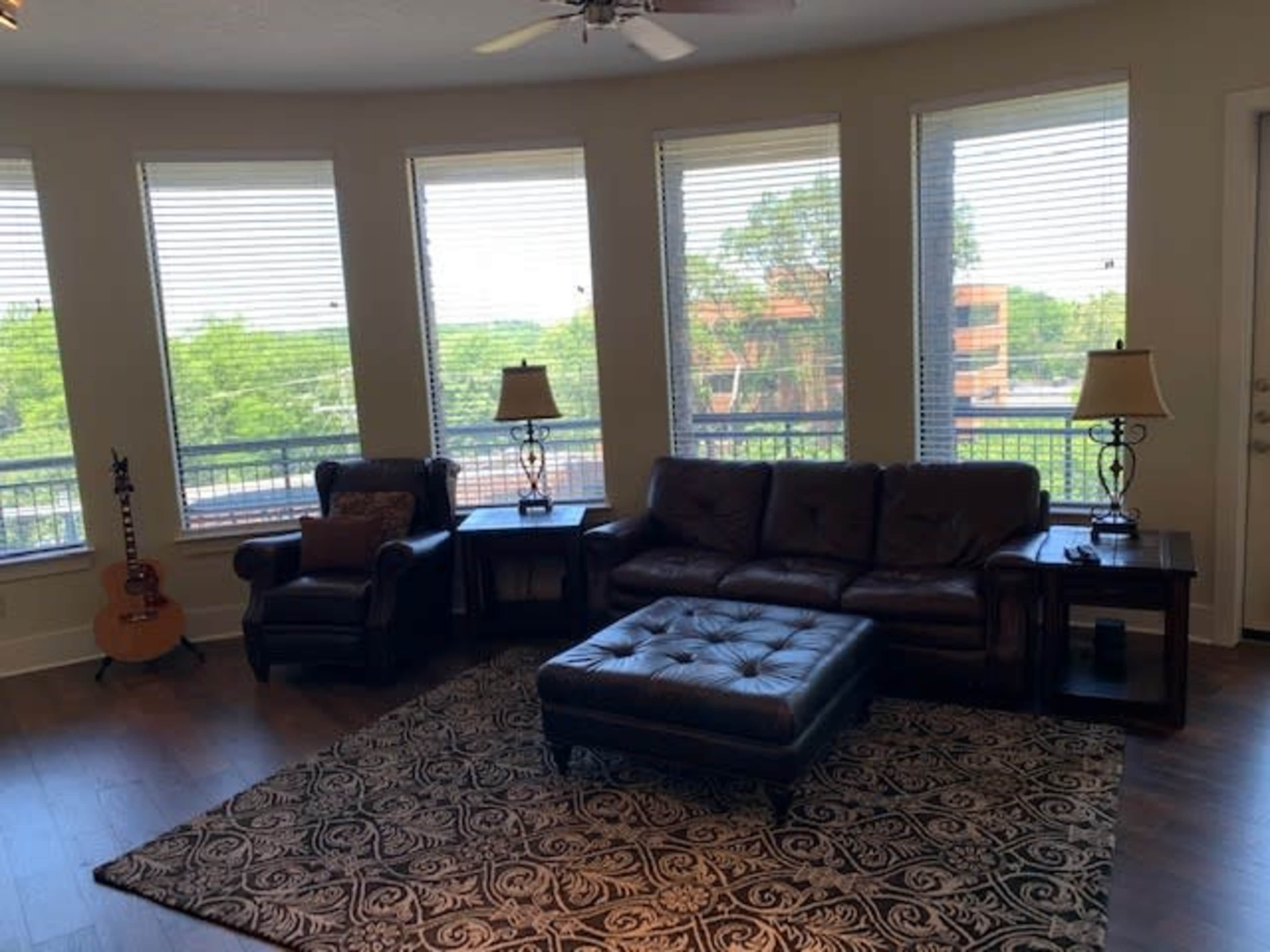 A cozy living room features a brown leather sofa, an armchair, a coffee table, and large windows offering a view of greenery outside.