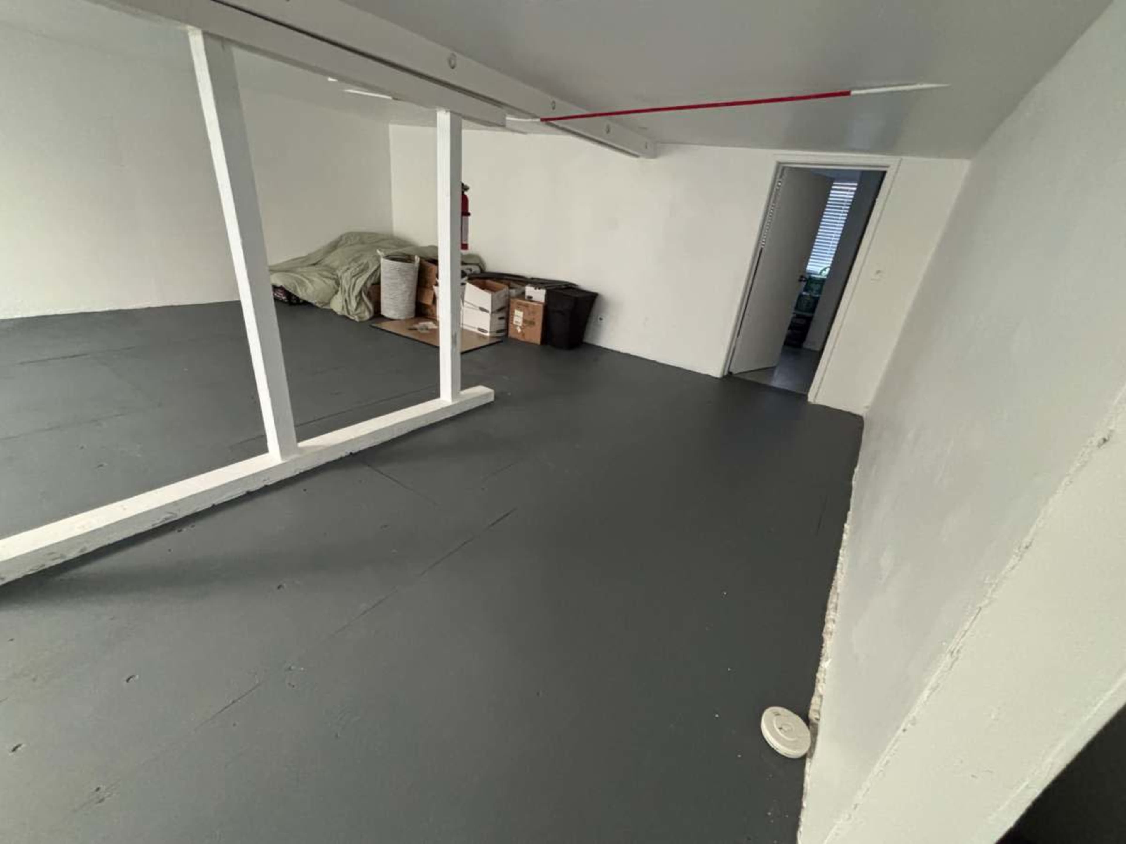 The image shows a sparsely furnished room with gray flooring, white walls, and a few cardboard boxes and items in the corner.