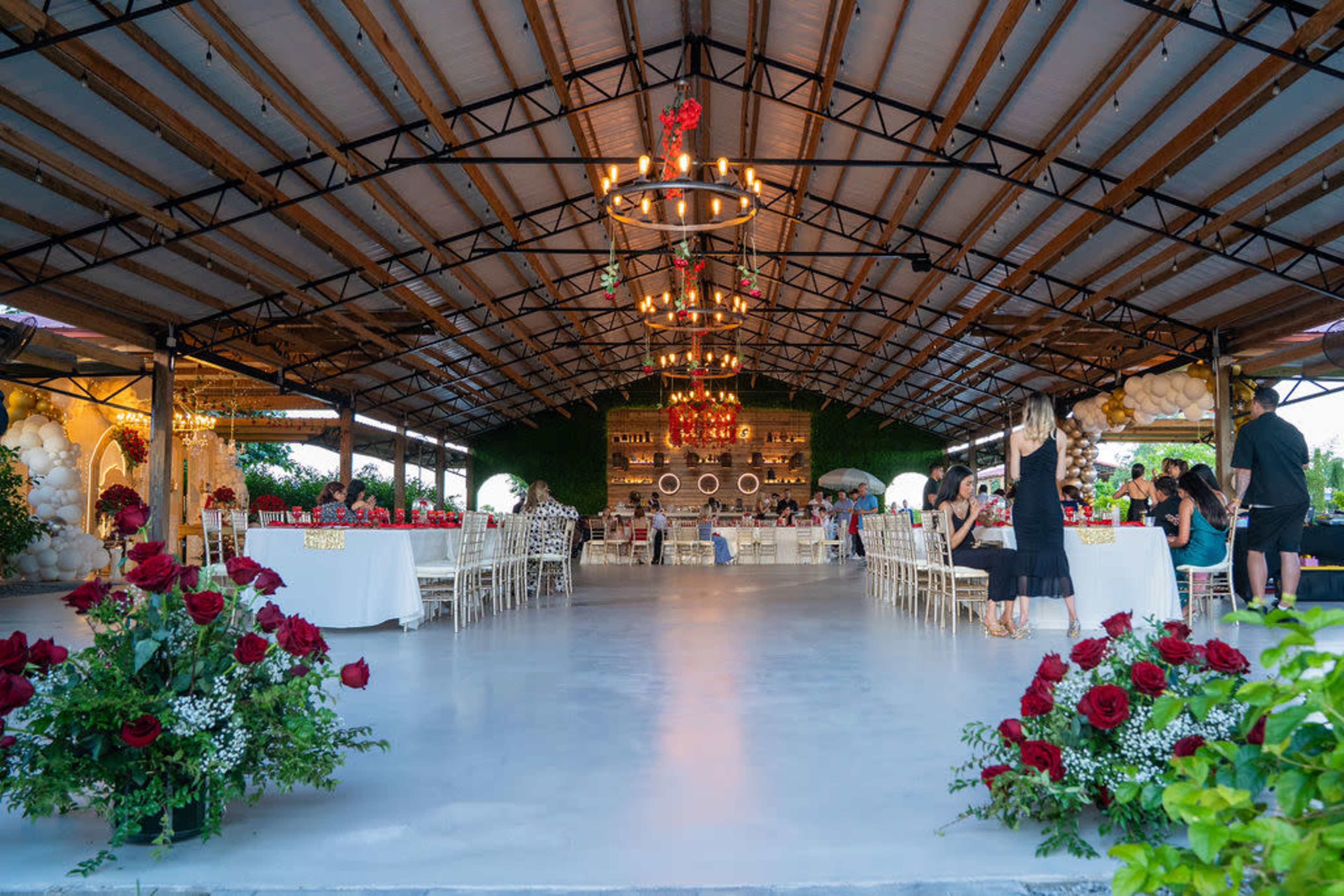 The image shows a spacious event venue decorated with tables and floral arrangements, featuring a large chandelier and a backdrop of greenery.