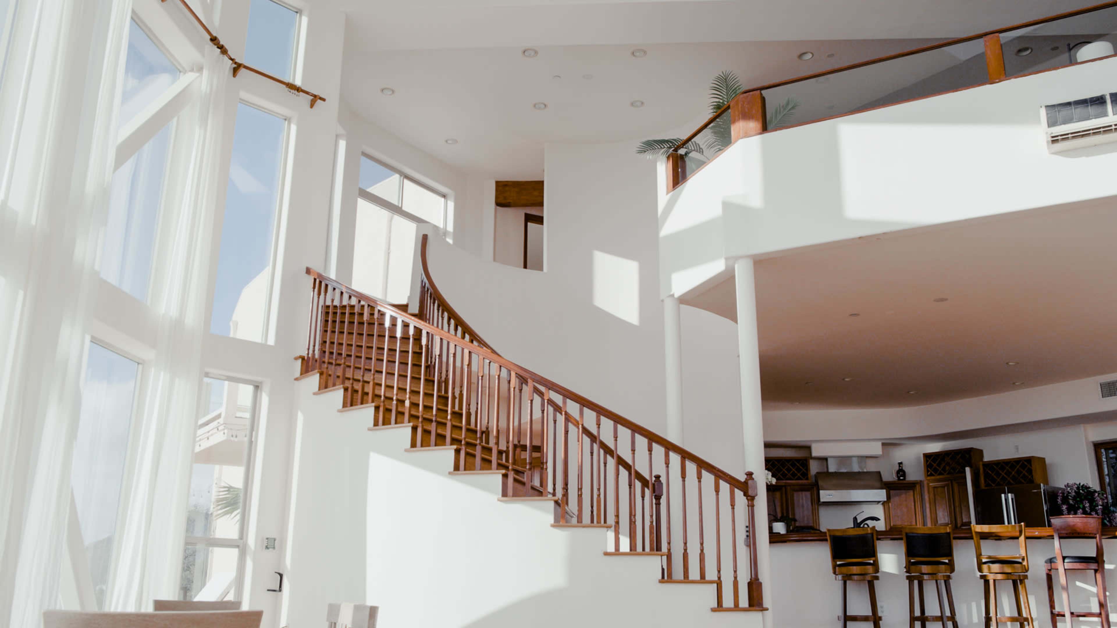 A spacious interior with a curved wooden staircase, large windows, and a bar area visible in the background.