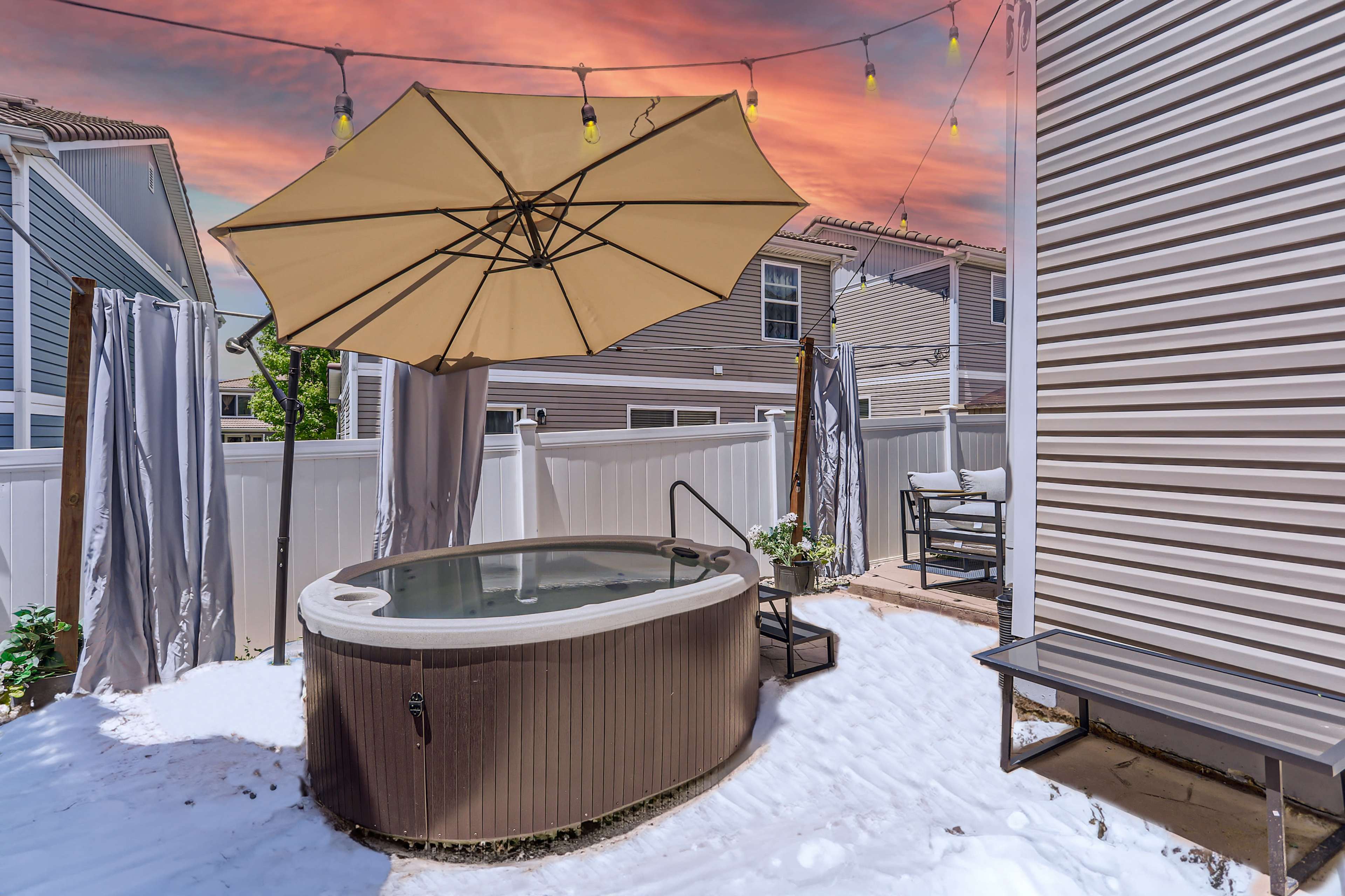 A hot tub is positioned beside a patio, surrounded by privacy curtains and an umbrella, under a colorful sky at sunset.