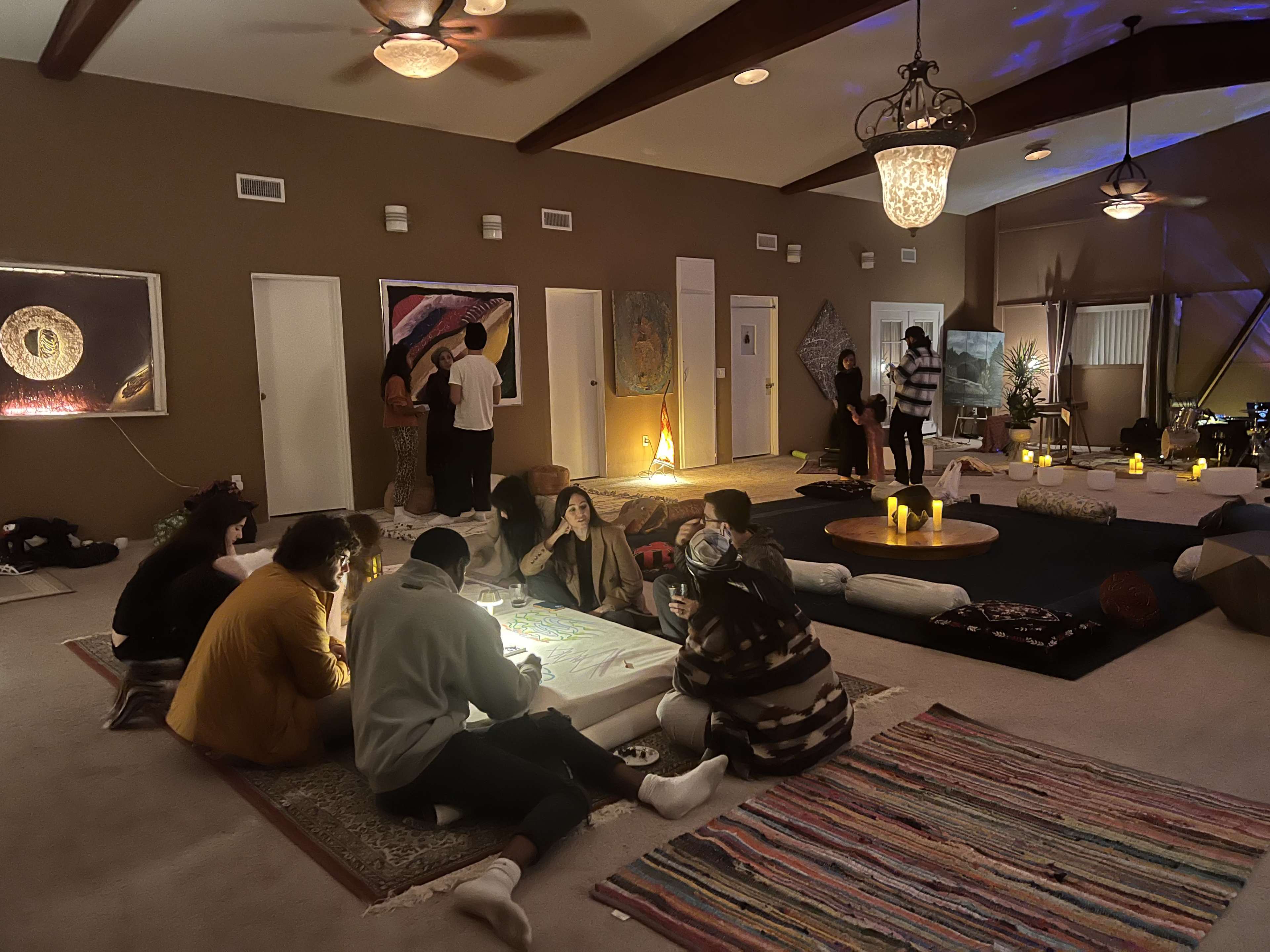 A group of people gathers in a spacious, softly lit room with art on the walls, candles, and floor cushions scattered around.
