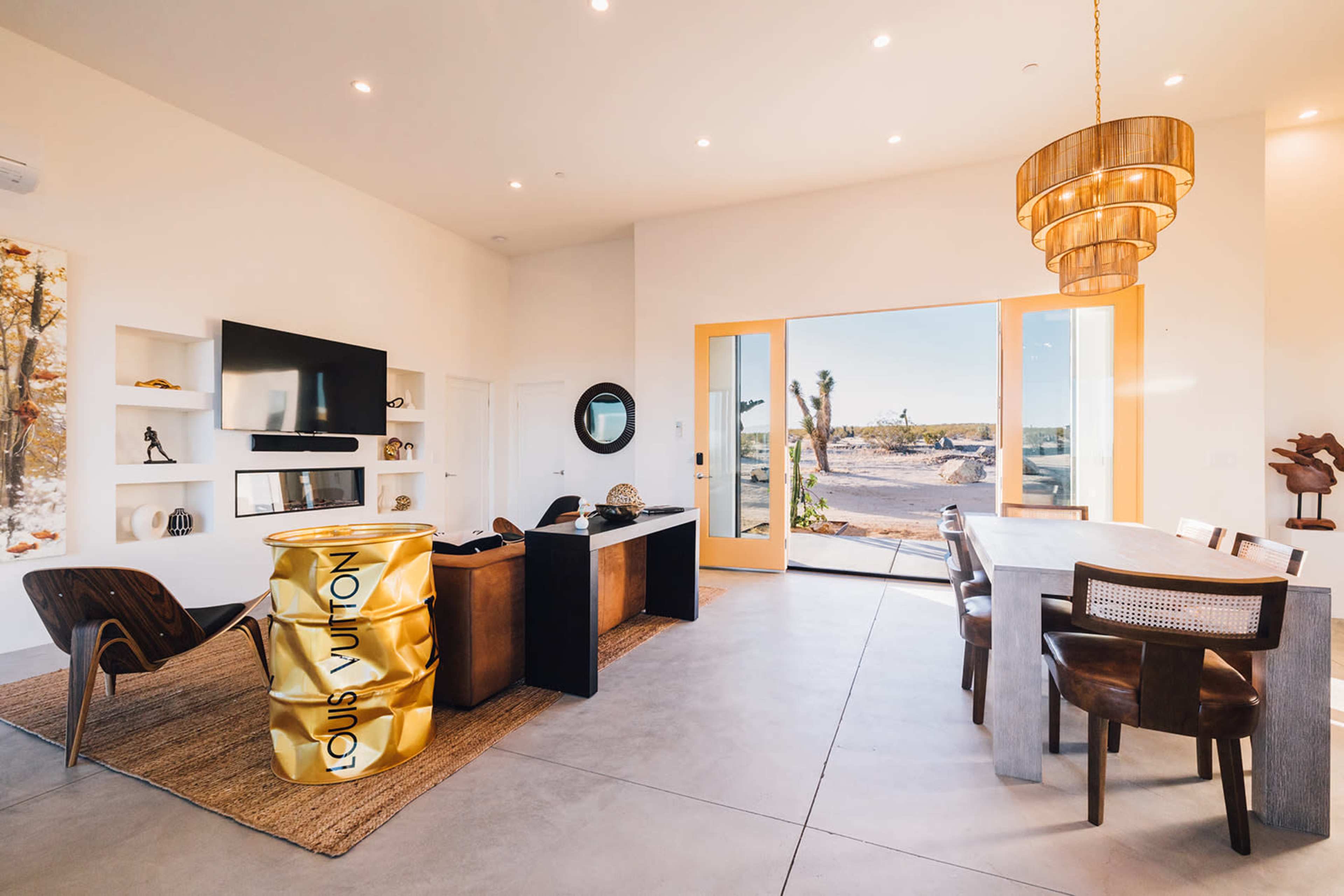 A modern interior space featuring a dining area with a large table, a sleek desk, and a view of the outdoor landscape through large glass doors.